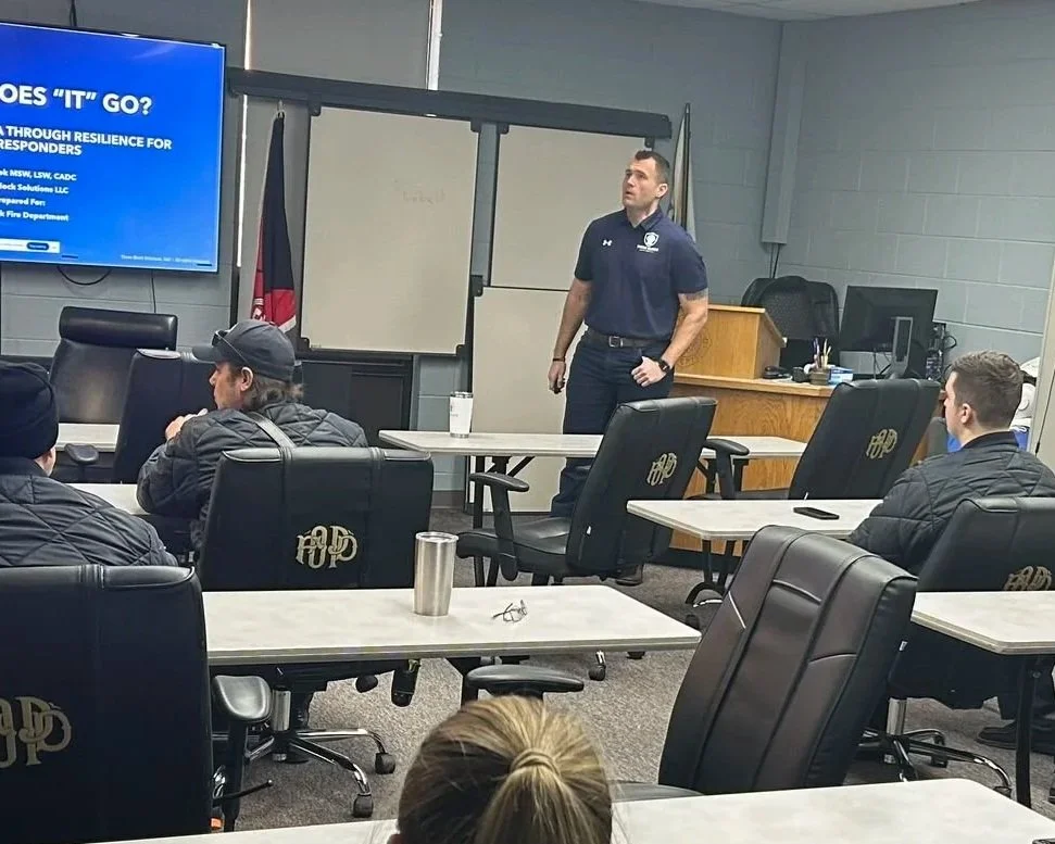 A classroom or training room with a man standing and giving a presentation to a group of seated attendees. The room has black chairs with gold logos, a large screen displaying a presentation slide, and a whiteboard. The attendees appear to be listening, some taking notes, with notebooks, laptops, and drinks on the tables.