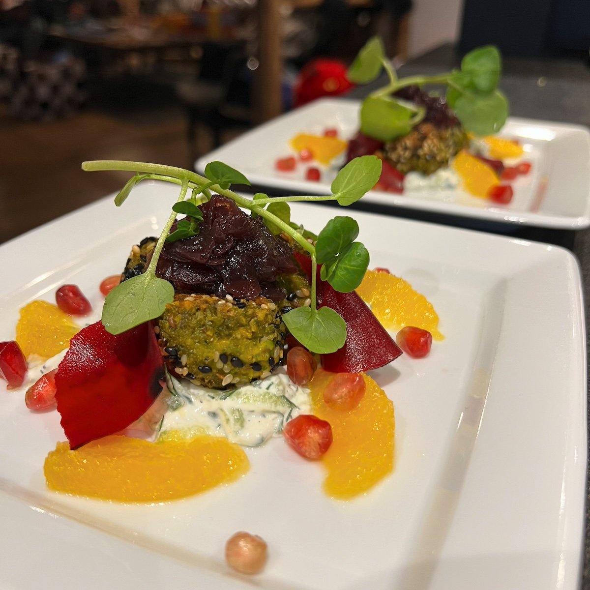 A plated gourmet dish with colorful ingredients including falafel, orange slices, pomegranate seeds, on a bed of tzatziki, garnished with microherbs.