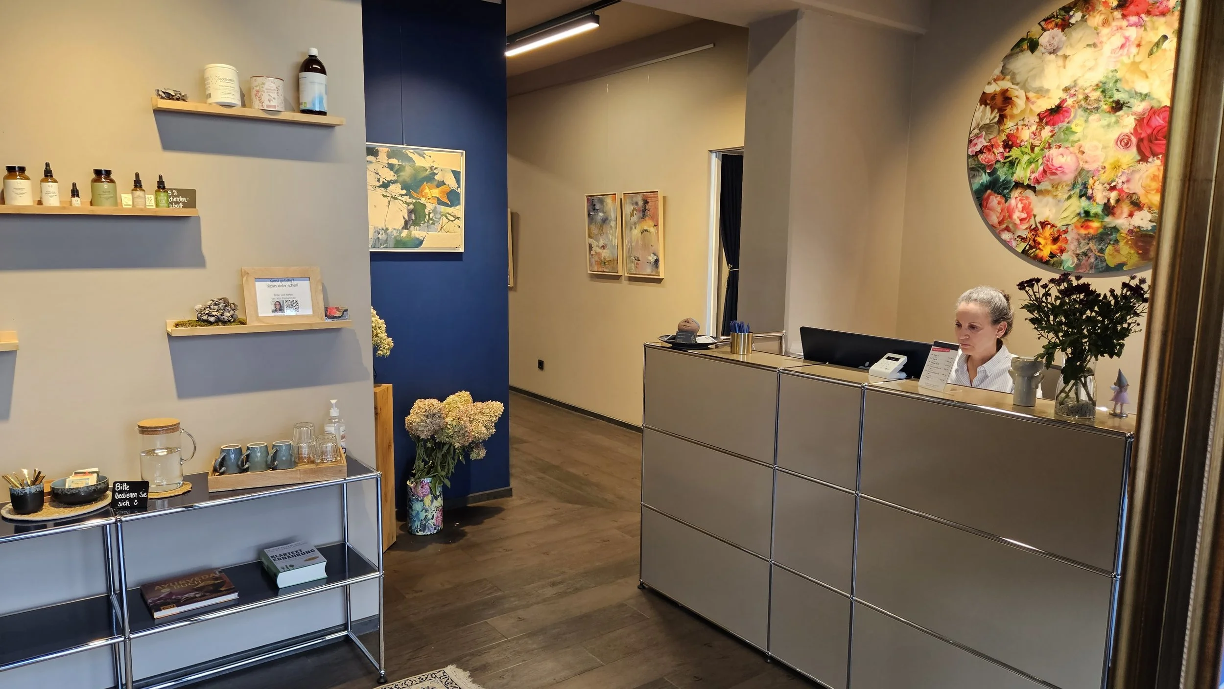 Rezeption eines Wellness- oder Spa-Bereichs mit einer erschwinglichen Mitarbeiterin hinter einem modernen Empfangstresen. Dekorative Blumen, bunte Wandkunst und Regale mit Kosmetik- und Pflegeprodukten.