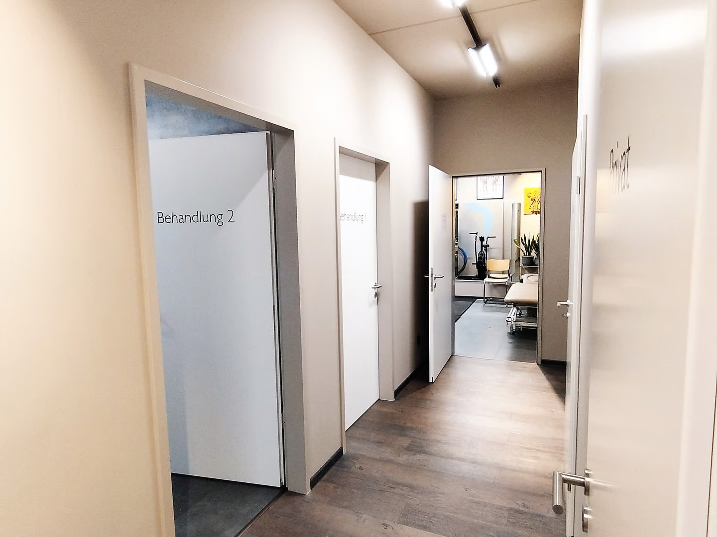 Ein Flur in einer medizinischen Einrichtung mit Türen zu Behandlungsräumen, darunter 'Behandlung 2' und andere Beschriftungen, sowie Wartezimmermöbel und Pflanzen im Hintergrund.