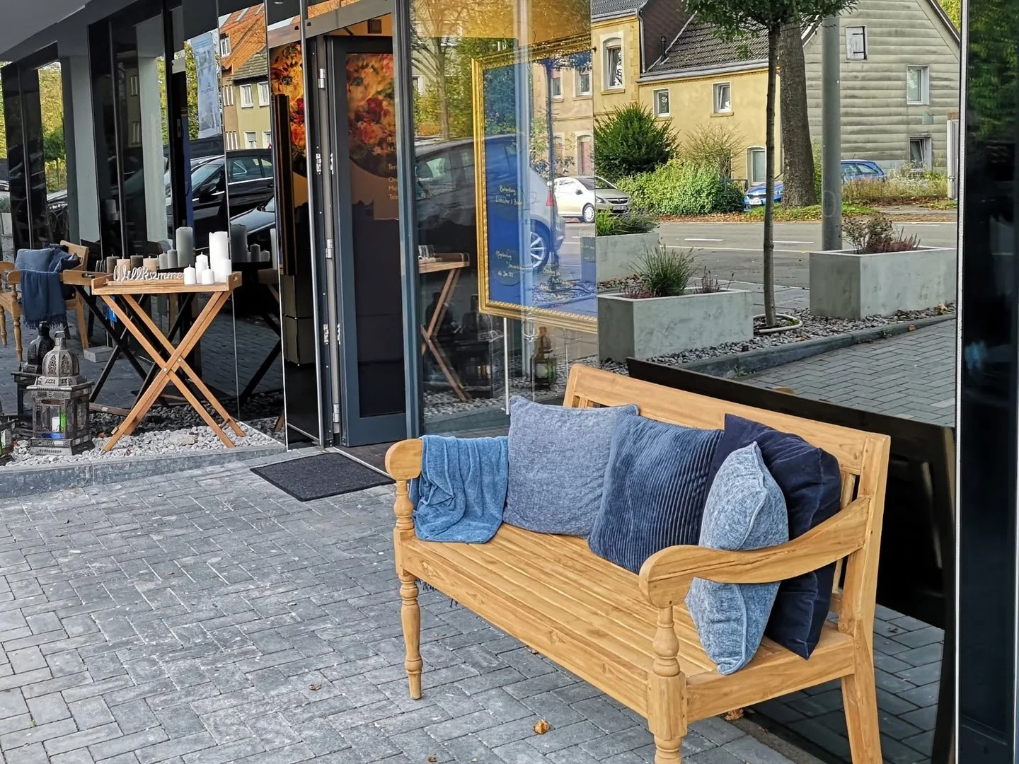Ein Laden mit Gartenmöbeln, darunter eine Holzbogenbank mit blauen Kissen, sowie eine Holzablage mit Kerzen und eine Laterne vor der Eingangstür, mit Blick auf die Straße und Häuser im Hintergrund.