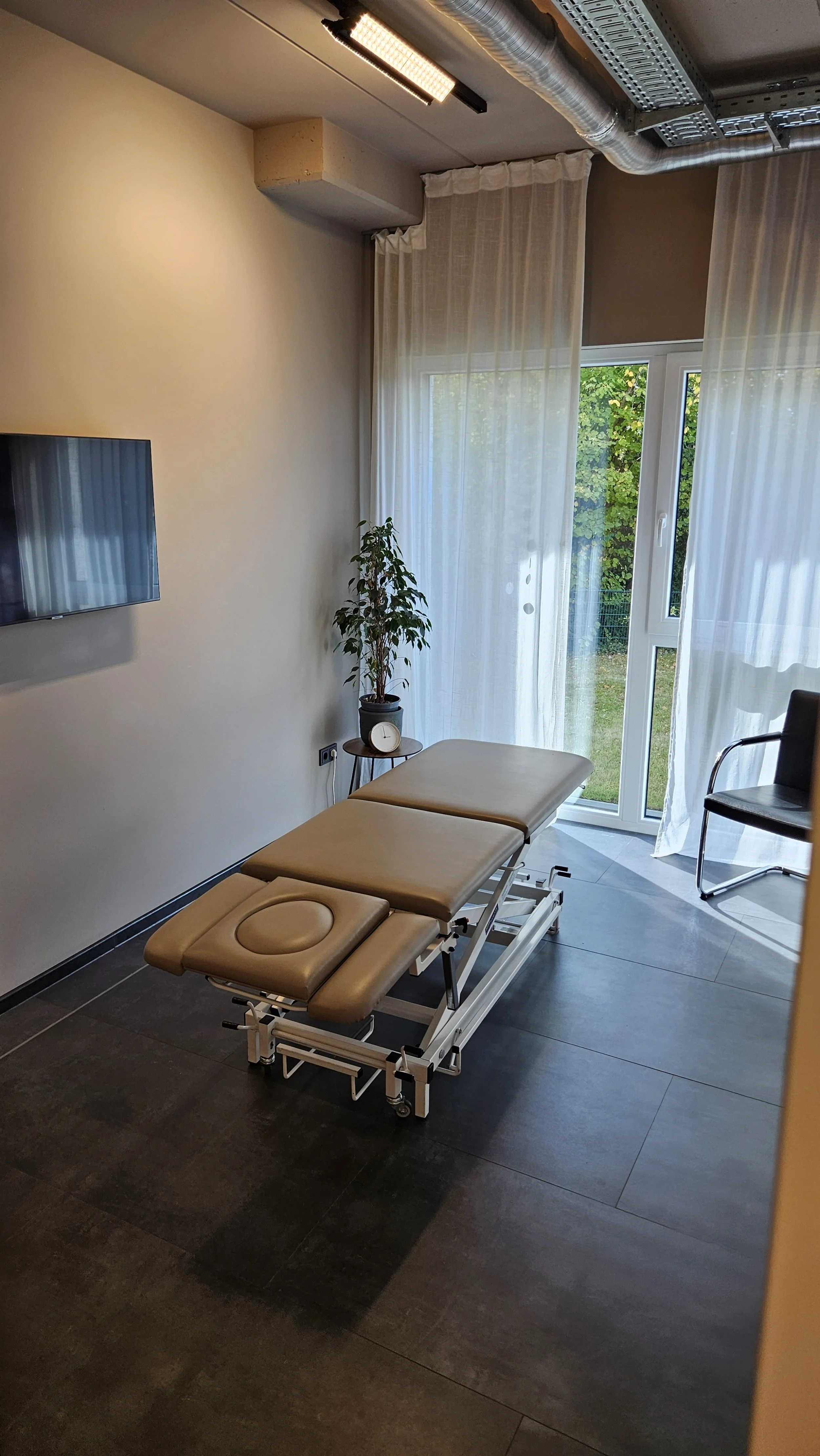 Ein Behandlungszimmer mit Behandlungsliege, einem Fernseher an der Wand, einem kleinen Tisch mit einer Pflanze und einer Uhr, sowie einem Stuhl, vor einem Fenster mit weißen Vorhängen.