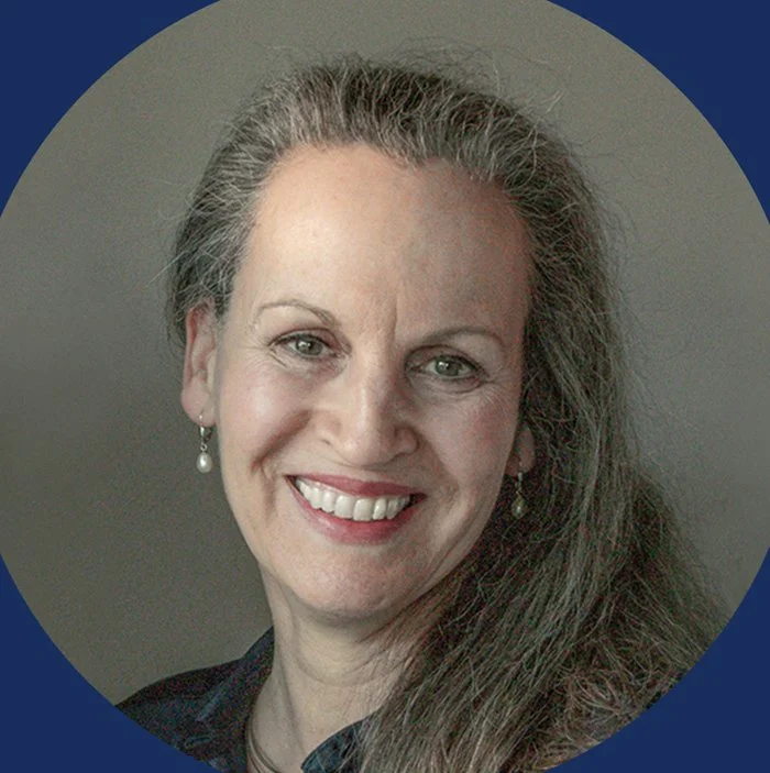 Ein Lächelndes Porträt einer Frau mit grauen Haaren, die Ohrringe trägt, vor einem neutralen Hintergrund.