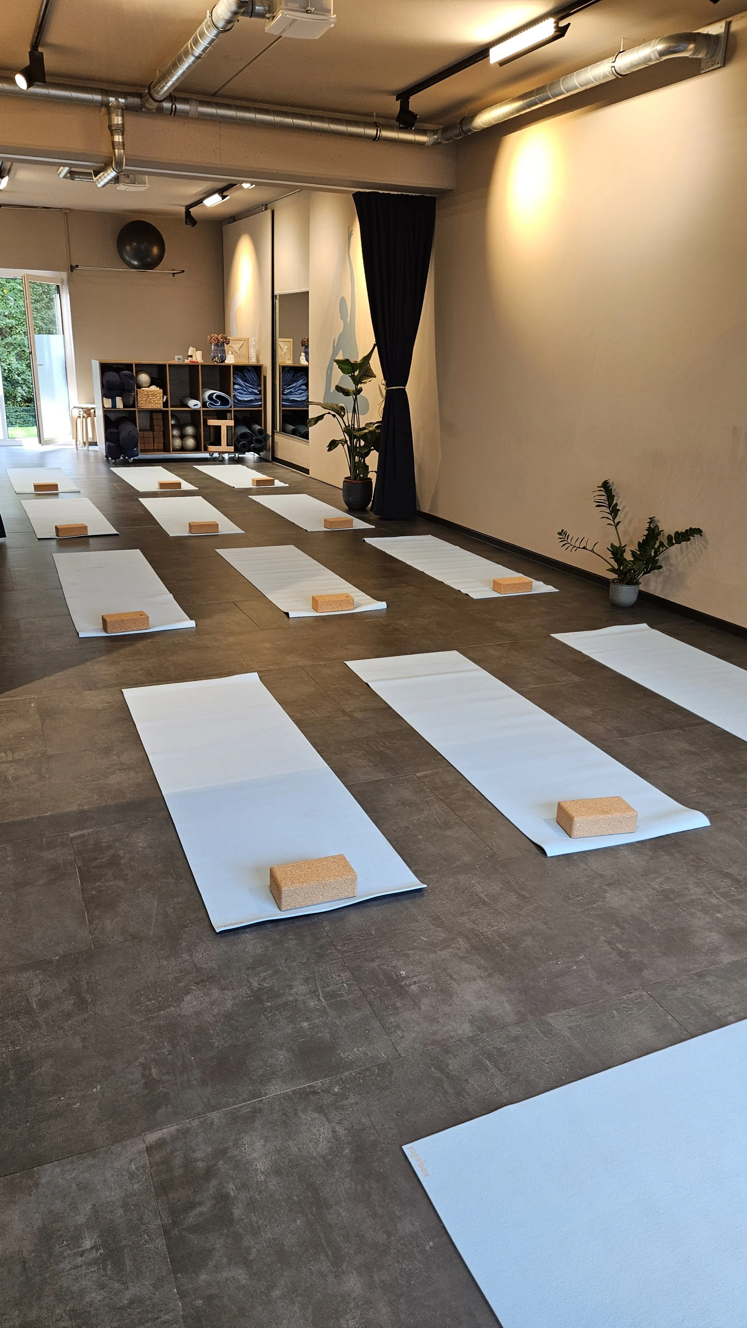Ein Yogaraum mit mehreren weißen Yogamatten, die mit Holzblöcken auf der Matte liegen. Im Hintergrund befinden sich Regale mit Yogazubehör, Pflanzen und ein Vorhang.