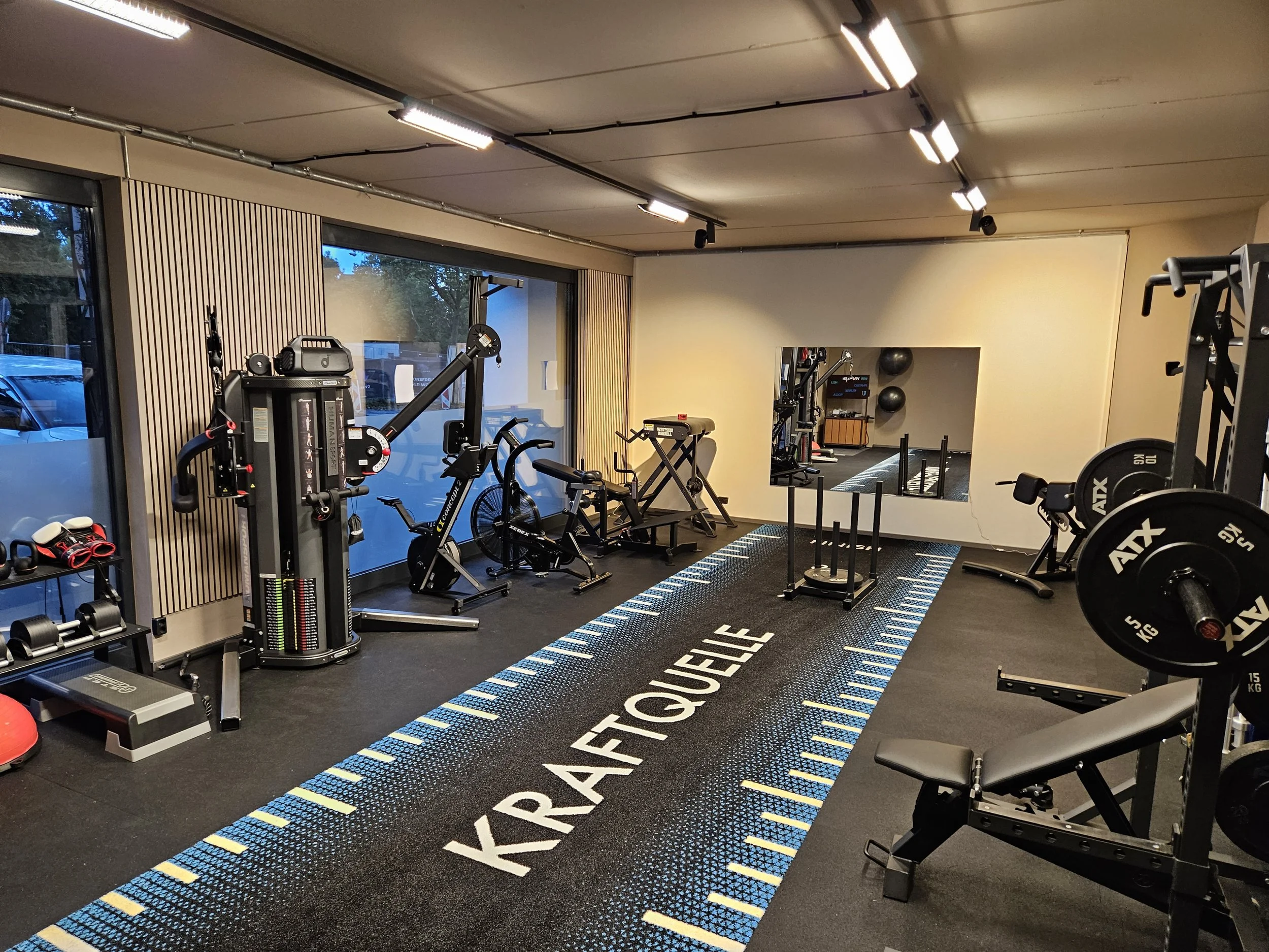 Innenraum eines Fitnessstudios mit verschiedenen Geräten wie Hanteln, Fahrrädern und Kraftgeräten, ein Spiegel an der Wand und ein Bodenmatte mit dem Schriftzug Krraftquelle.