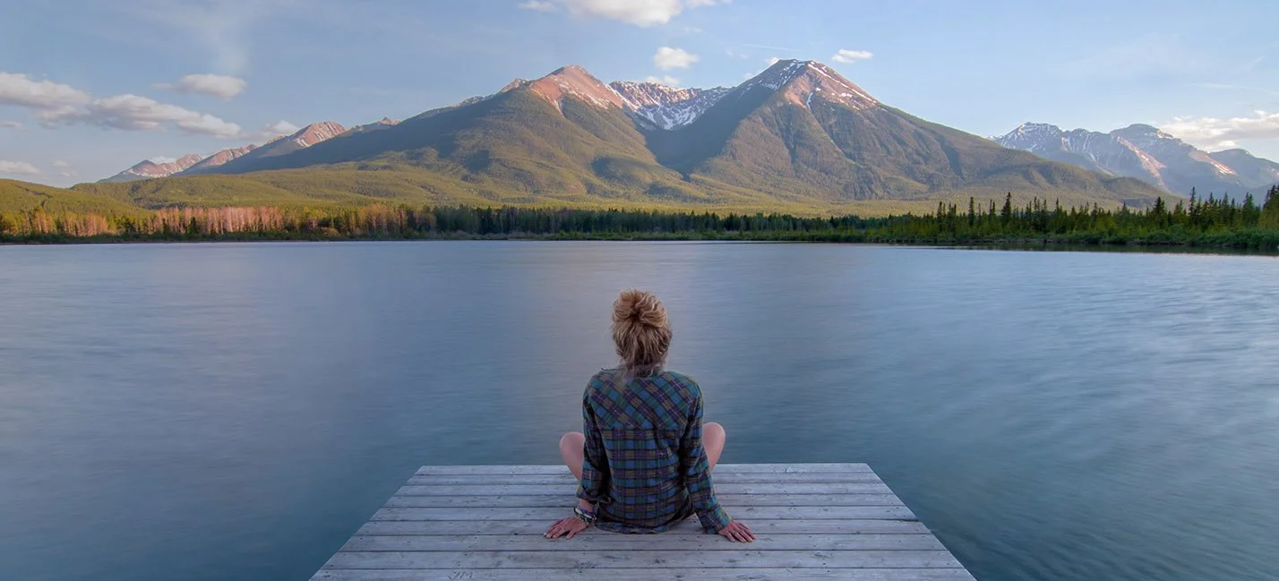 Person sitzt auf einem Steg am Wasser, blickt auf Berge im Hintergrund.