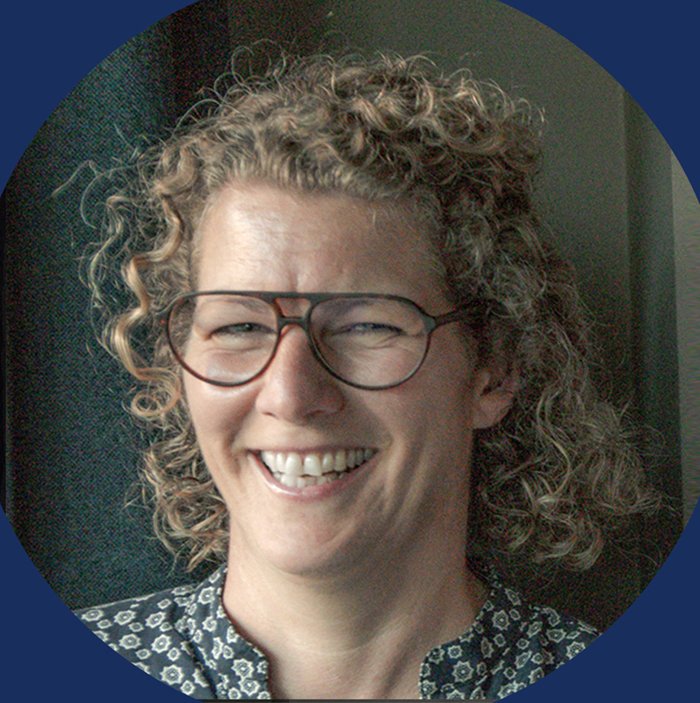 Lächelnde Frau mit Brille und lockigem Haar, im Hintergrund eine dunkle Wand.