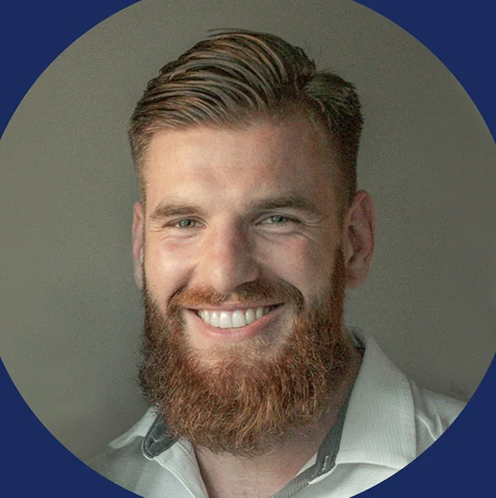 Ein freundlicher Mann mit braunem Bart und blondem Haar, trägt ein weißes Hemd, lächelt in die Kamera.