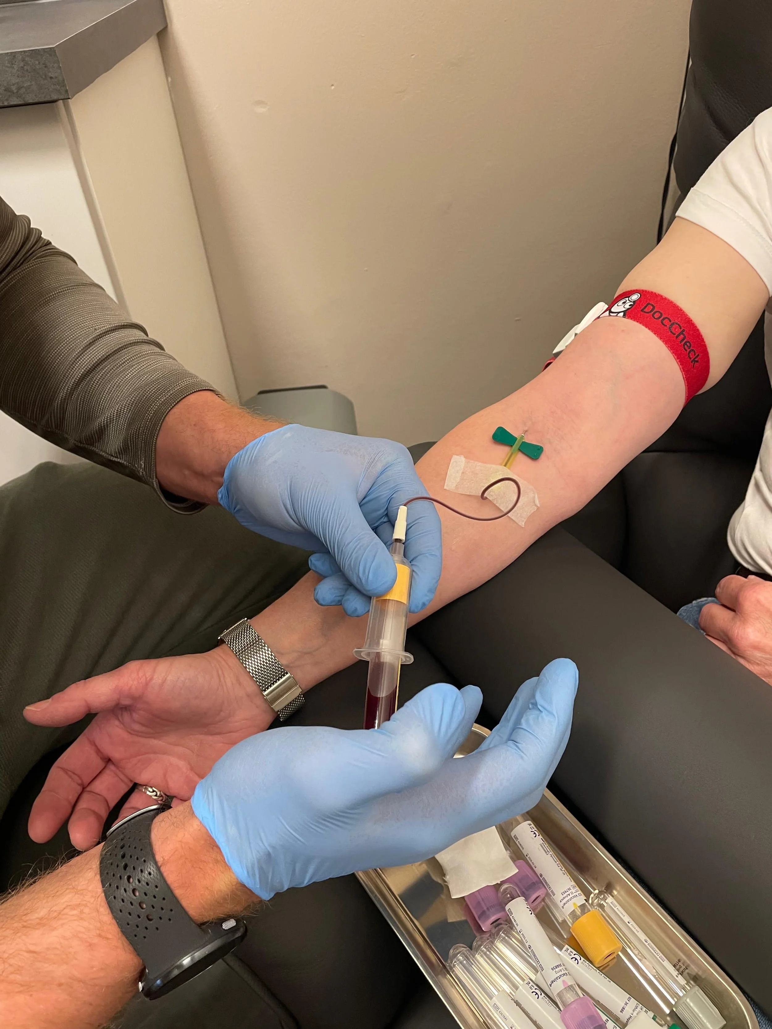 Ein Arzt entnimmt Blut aus einem Arm eines Patienten, der auf einer Liege sitzt. Der Patient trägt einen roten Armband mit der Aufschrift 'DocCheck'. Die medizinische Ausrüstung liegt auf einem Tablett.