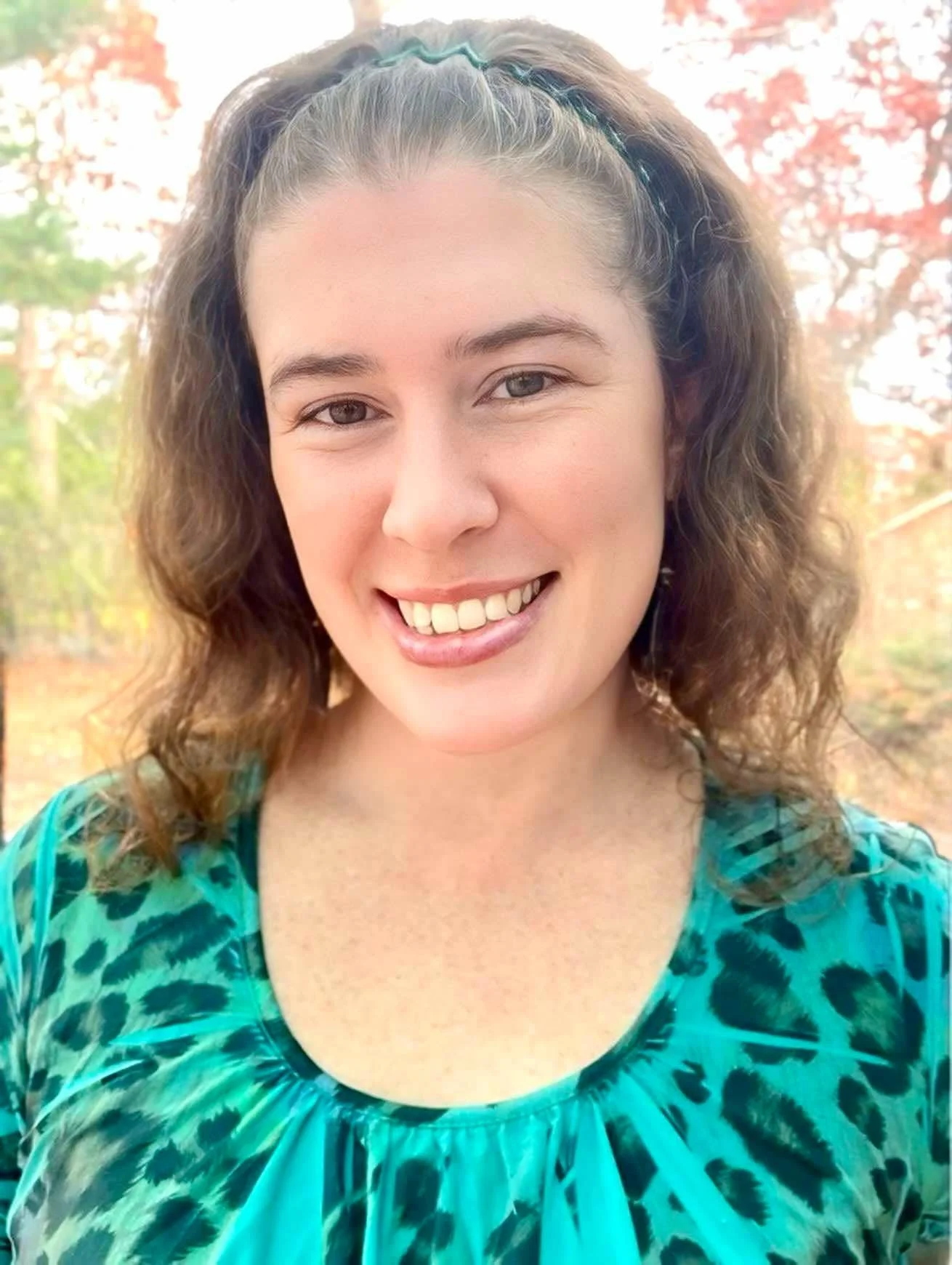 A young woman with curly brown hair, wearing a teal and black animal print top, smiling outdoors with trees and autumn leaves in the background.