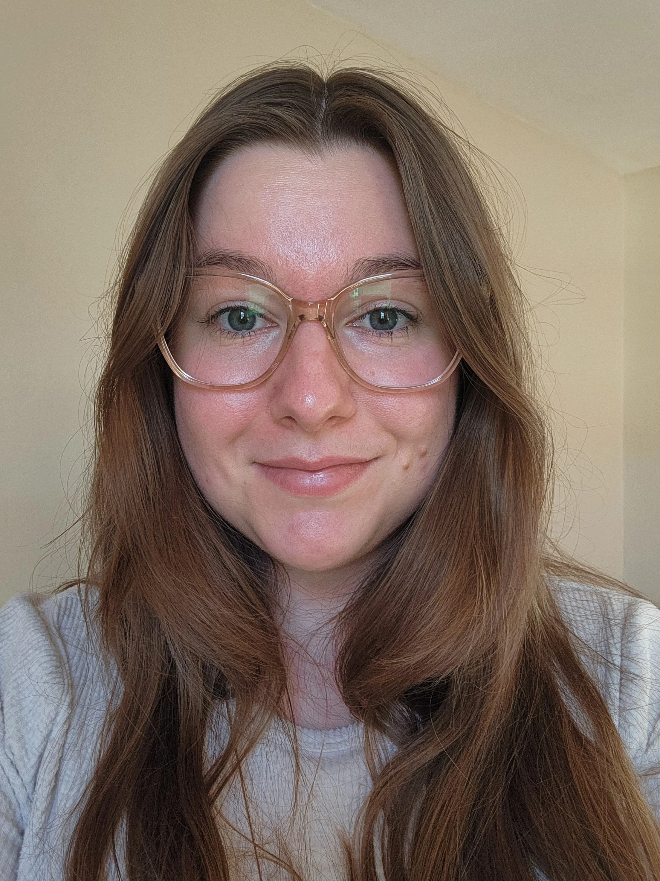 A woman with long, wavy brown hair wearing clear glasses and a white top, smiling slightly in front of a beige wall.