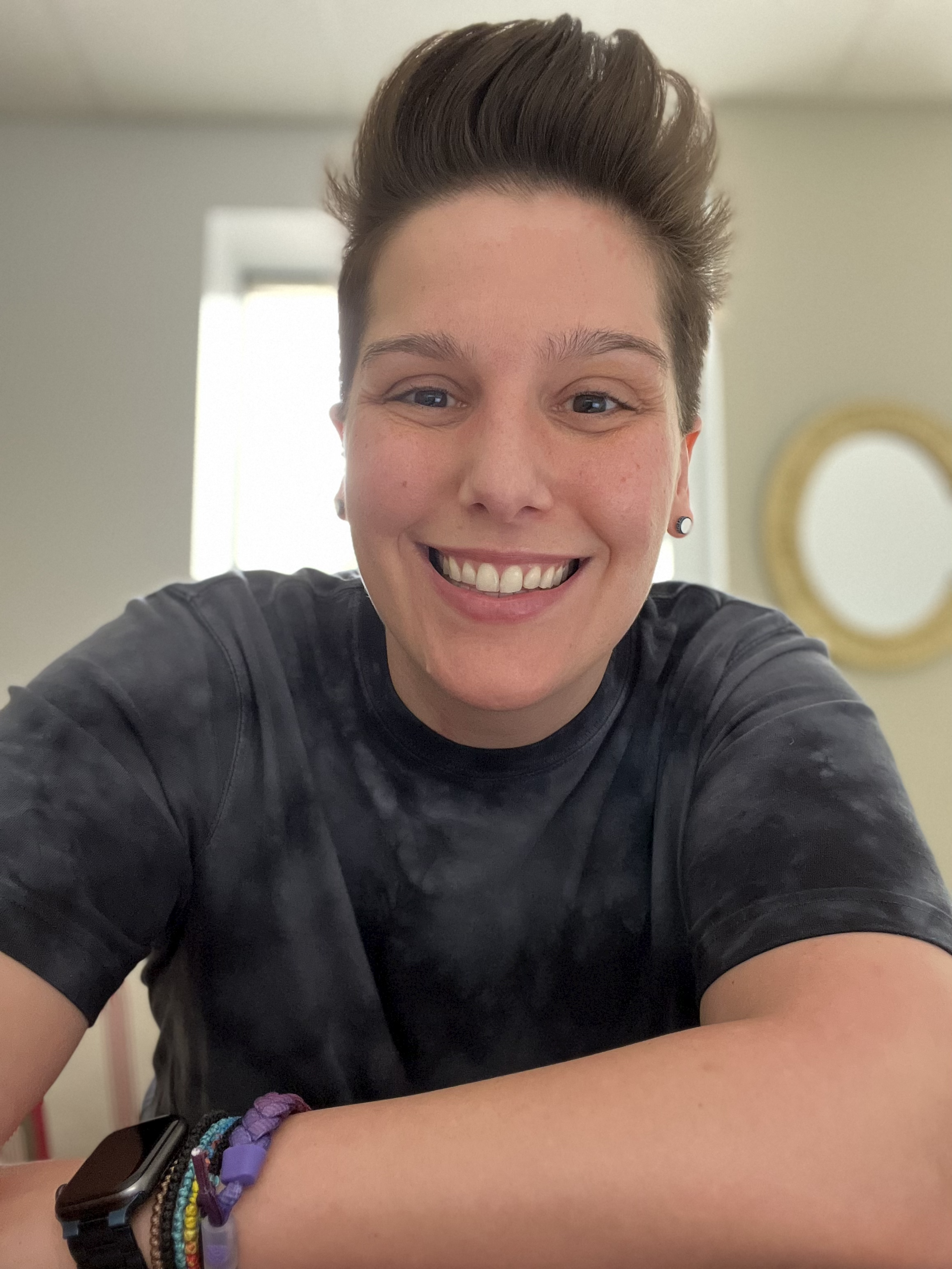 Close-up of a young woman with short styled hair, smiling at the camera, wearing a black shirt, a black watch, and colorful bracelets on her wrist, with a mirror and window in the background.