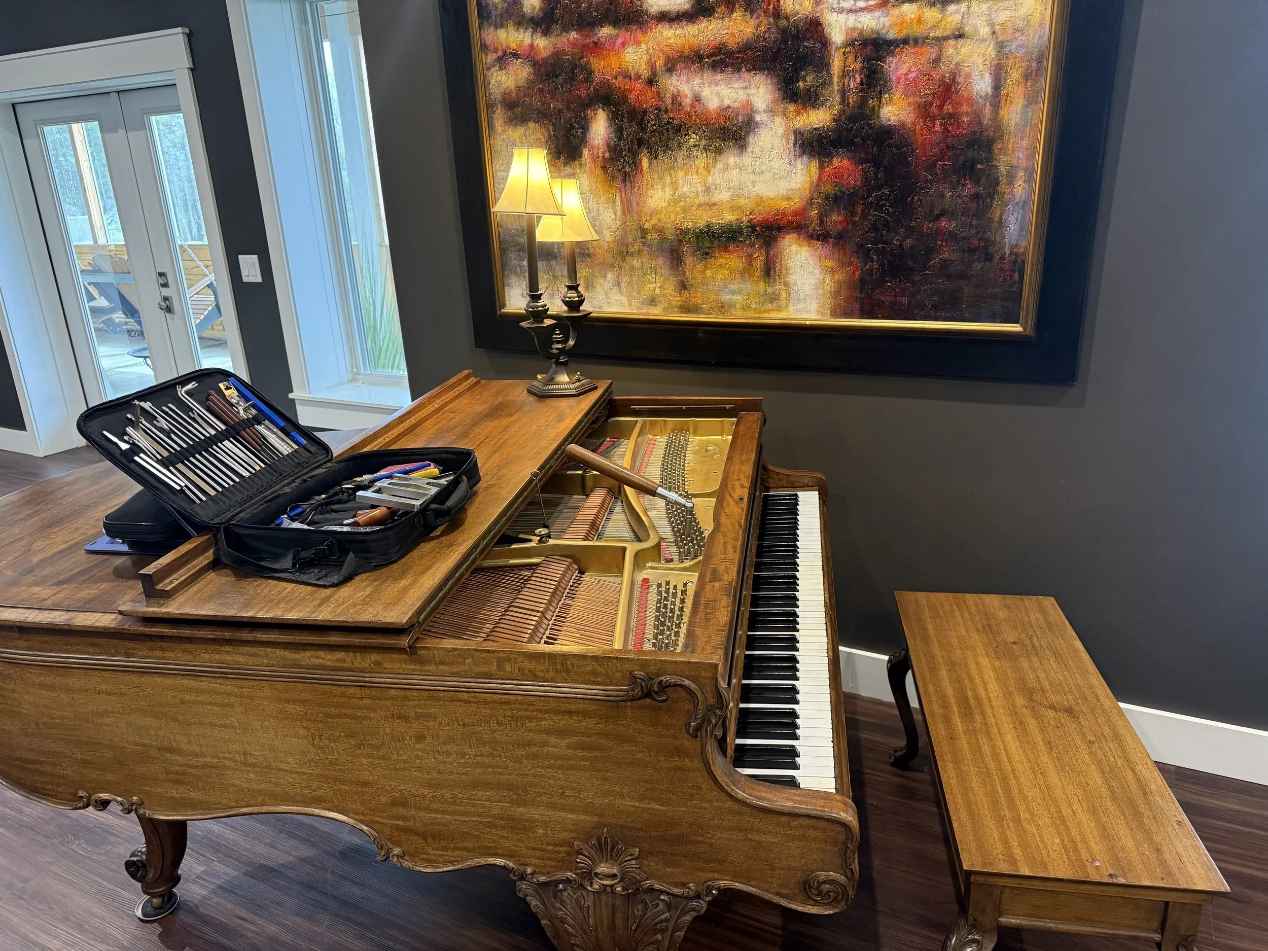An open vintage wooden grand piano with the lid raised, situated in a room with dark gray walls and hardwood flooring. A small matching wooden bench is positioned in front of the keyboard. On top of the piano is a black tool kit containing various mu