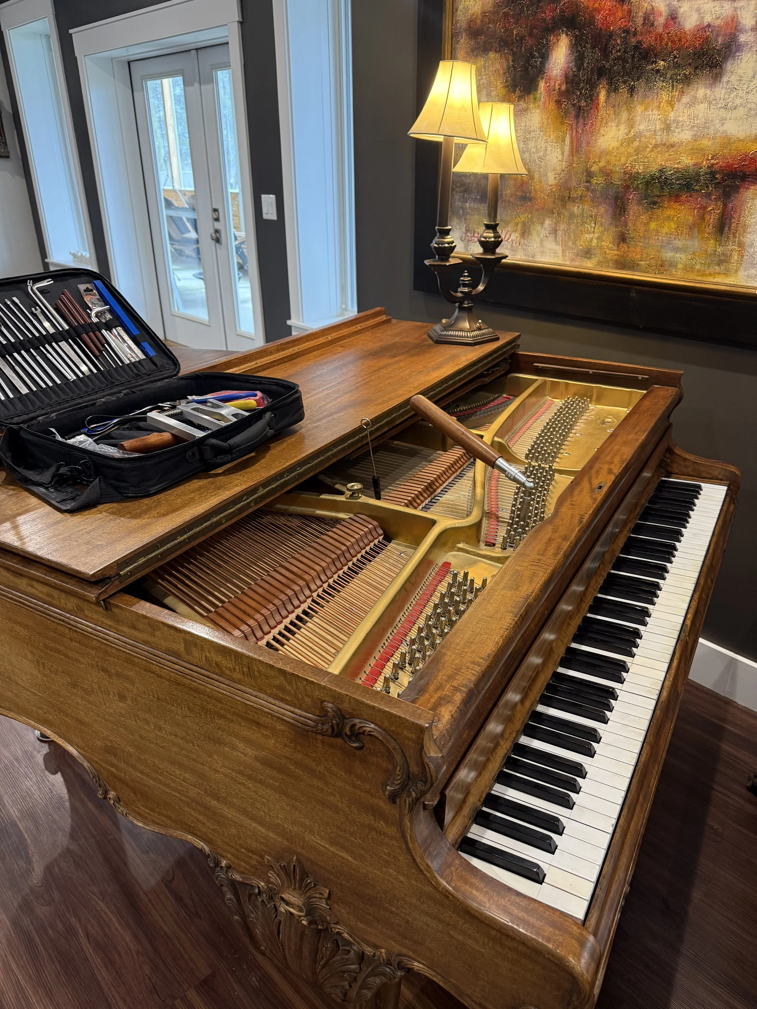 A wooden upright piano with the top open, revealing its strings and hammers. There is a tuning tool and a case of piano tuning tools resting on the lid. Behind the piano, there is a lamp with two shades and a colorful abstract painting on the wall. I