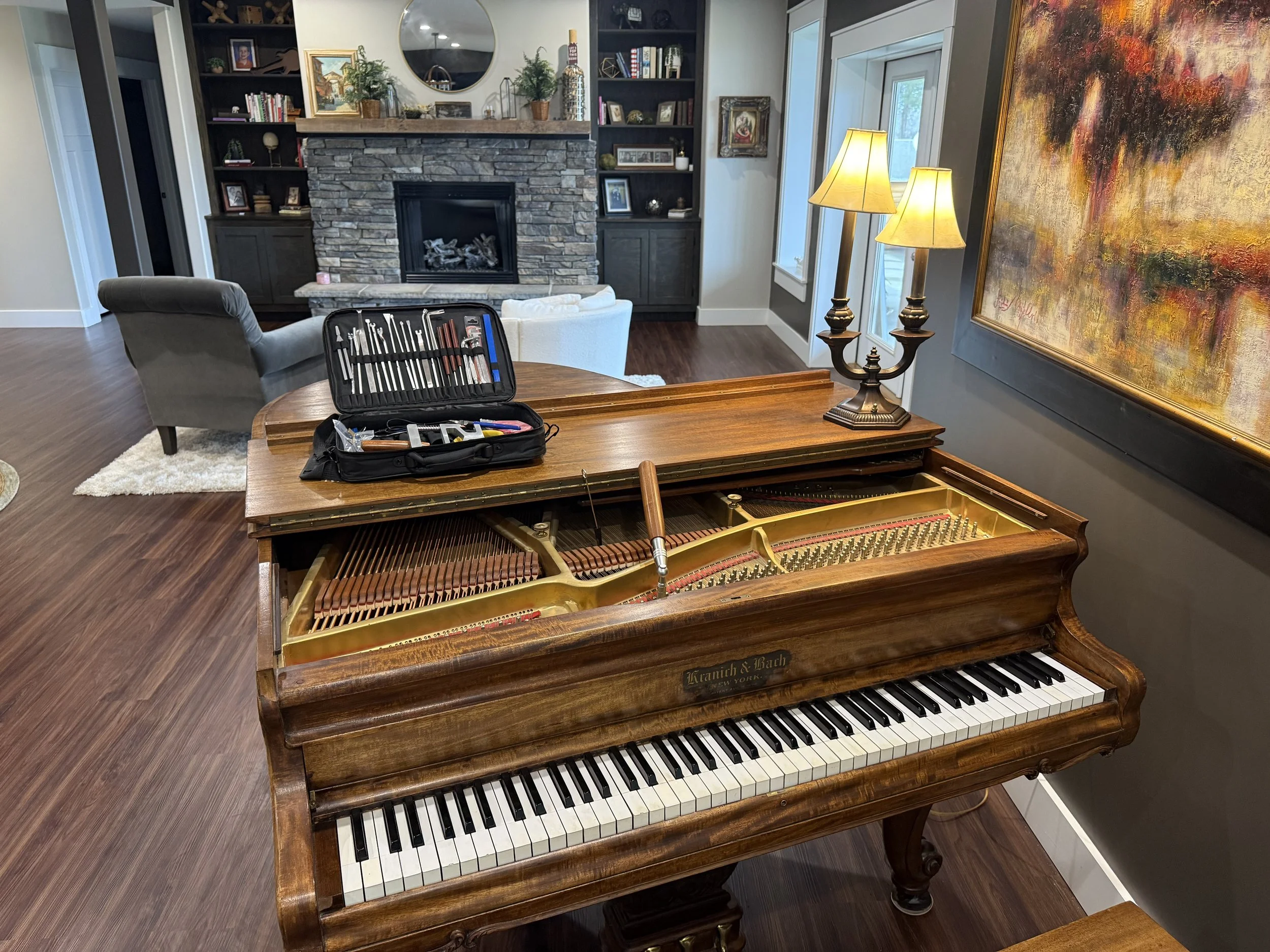 A vintage brown piano with a music stand and a repair kit on top, situated in a cozy living room with a fireplace, built-in shelves, a grey armchair, and a painting, near a window with a pair of lamps.