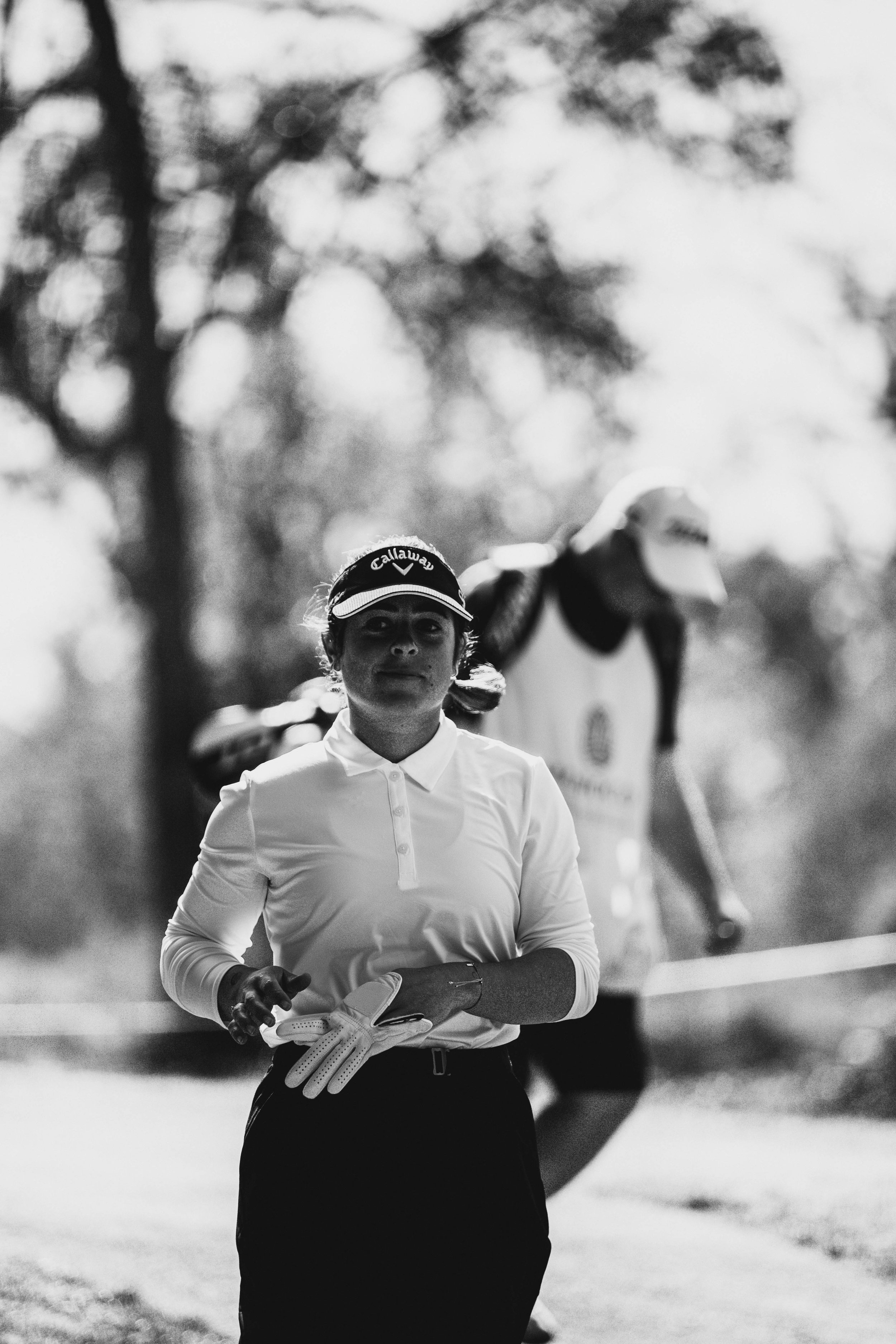 Persona preparándose para jugar golf, en blanco y negro. Lleva una visera y guantes de golf. Al fondo, otra persona con uniforme de caddie.