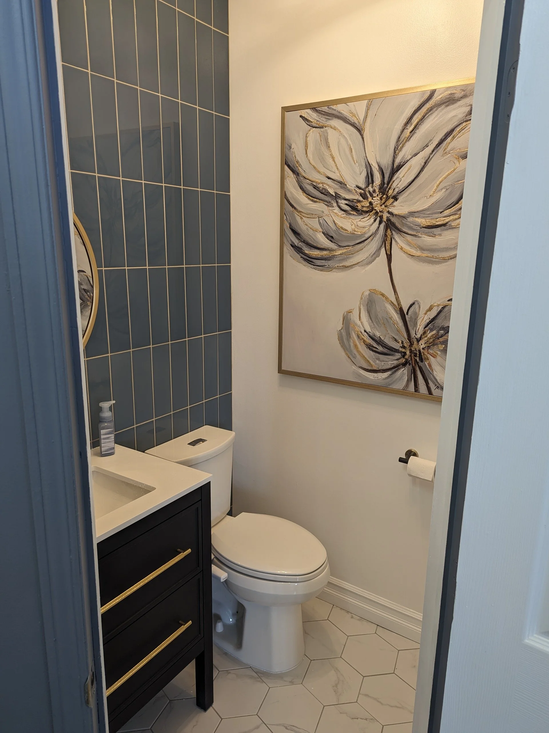 Modern bathroom with tiled walls, a black vanity with gold handles, white toilet, hexagonal floor tiles, hand soap dispenser, and floral wall art.