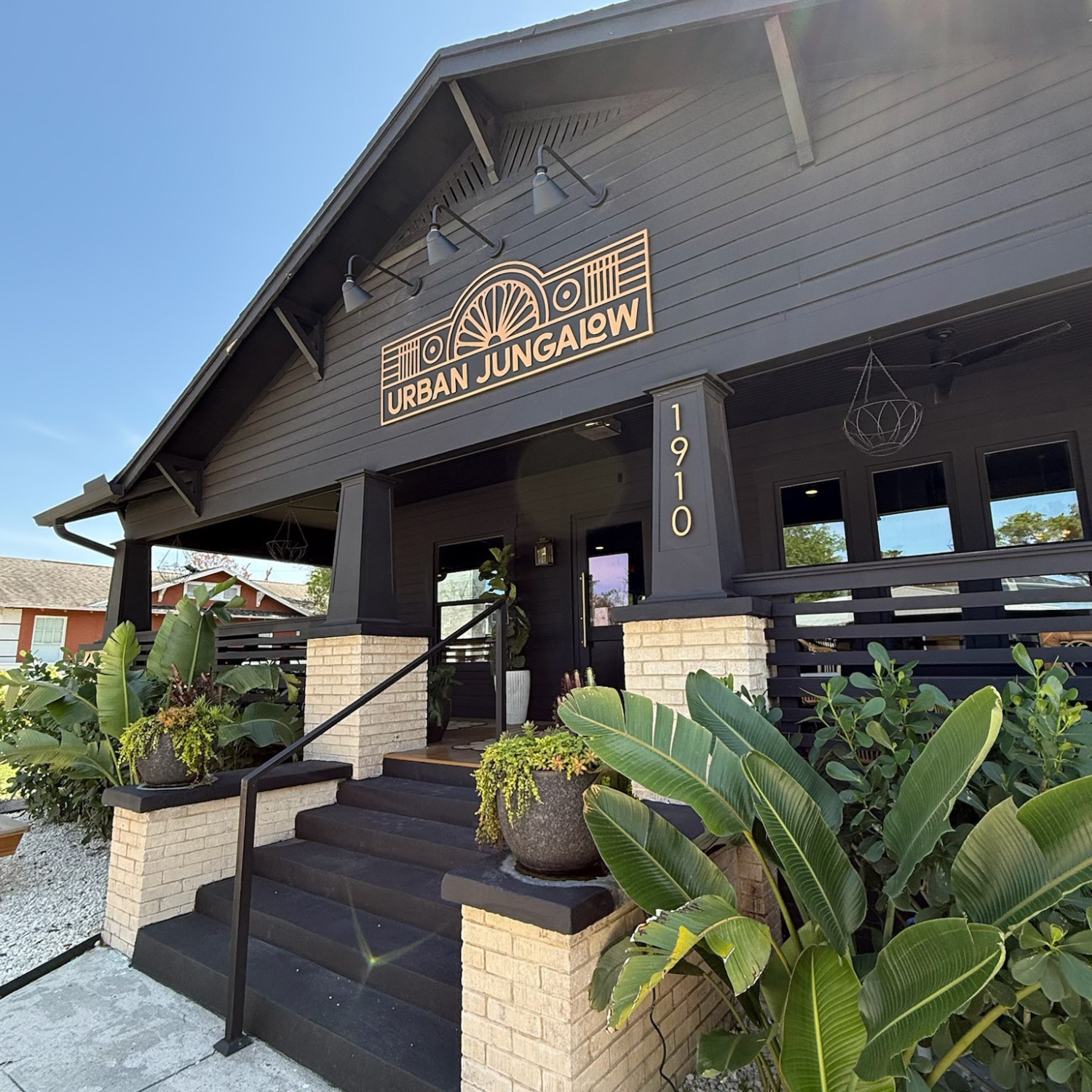 The exterior of a black building with a sign that reads 'Urban Jungalow' number 1910, surrounded by tropical plants and greenery.