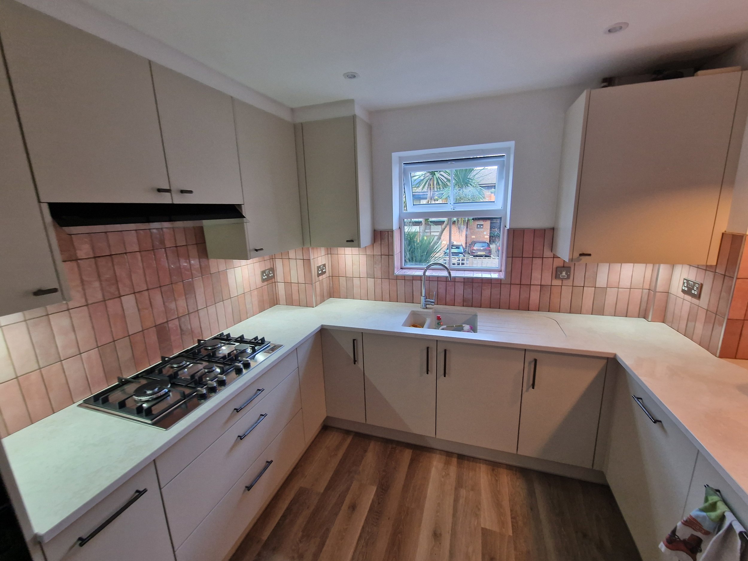 tile installation experts Modern kitchen with white cabinets, sleek pink tile backsplash, gas stove, and sink under a window. kitchen tiled by tile infinity in winchester, uk