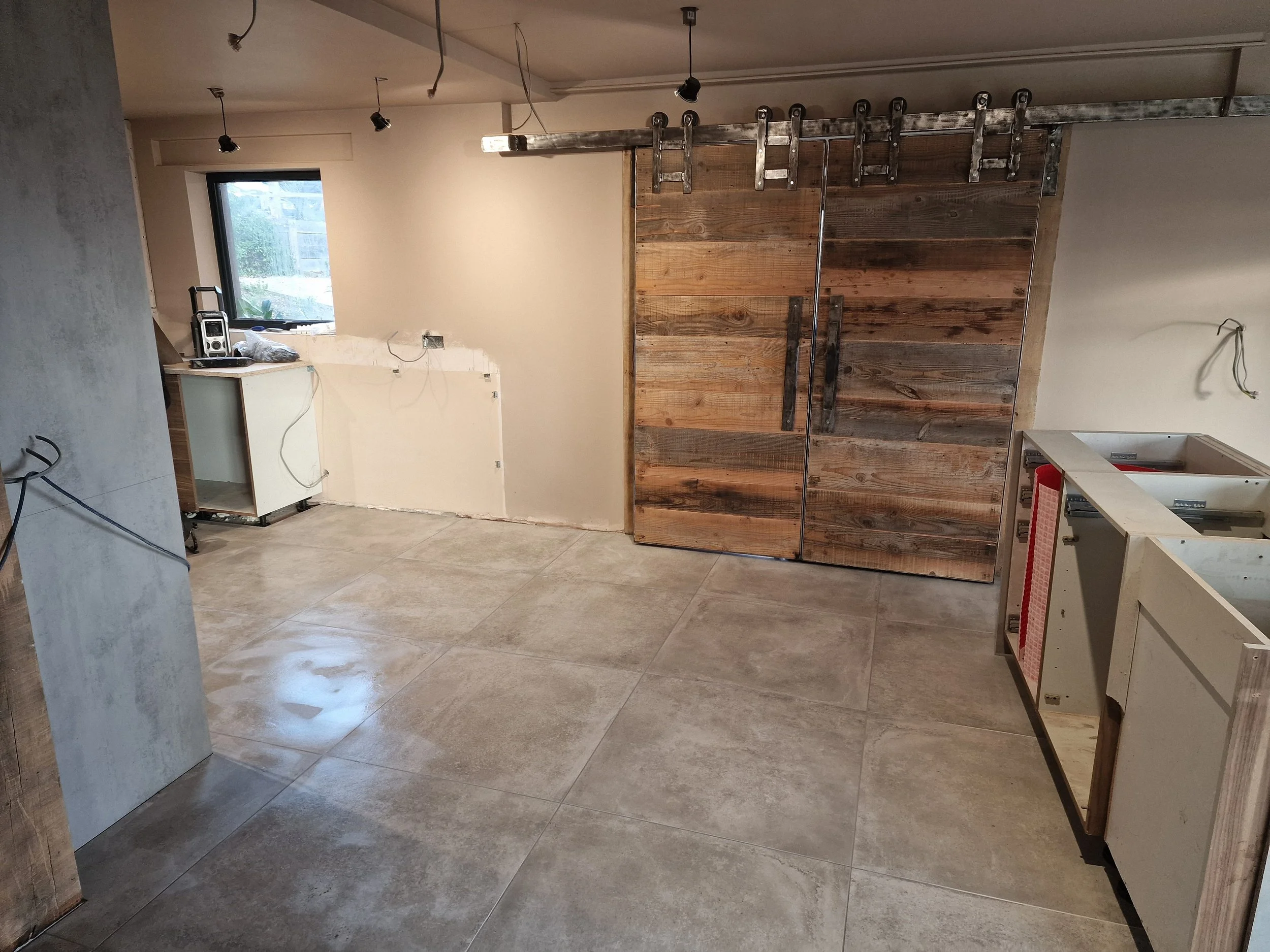 Interior of a barn room under renovation with unfinished walls, tile flooring, and a rustic wooden sliding door. Partially installed cabinets and visible wiring indicate ongoing construction work. New kitchen tiling project in Southampton" fareham