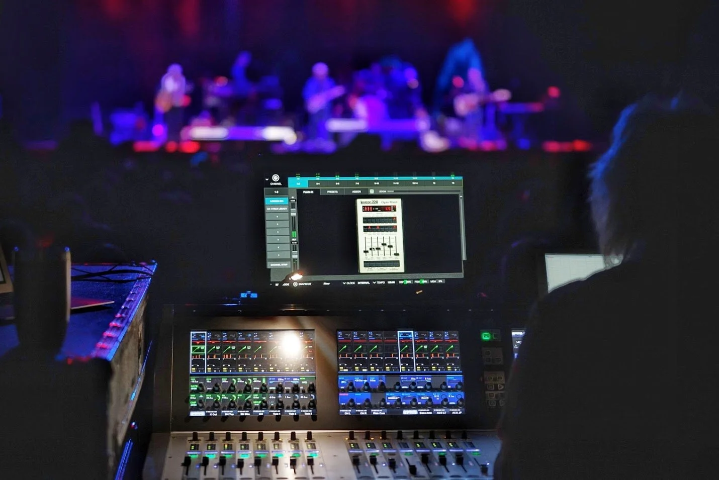 A sound engineer working at a mixing console during a live concert with a band playing on stage in the background.