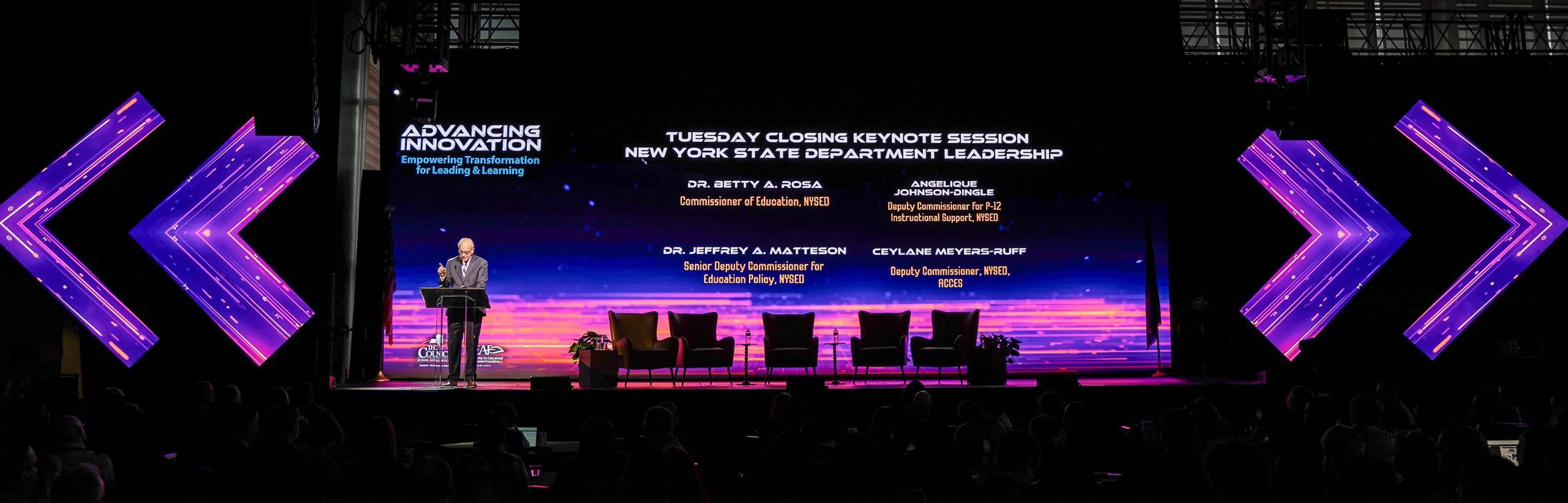 Panel discussion stage at a conference with a large screen displaying speaker names and titles. A man in a suit is speaking at a podium, and five empty chairs are arranged on stage. Audience members are seated in front.