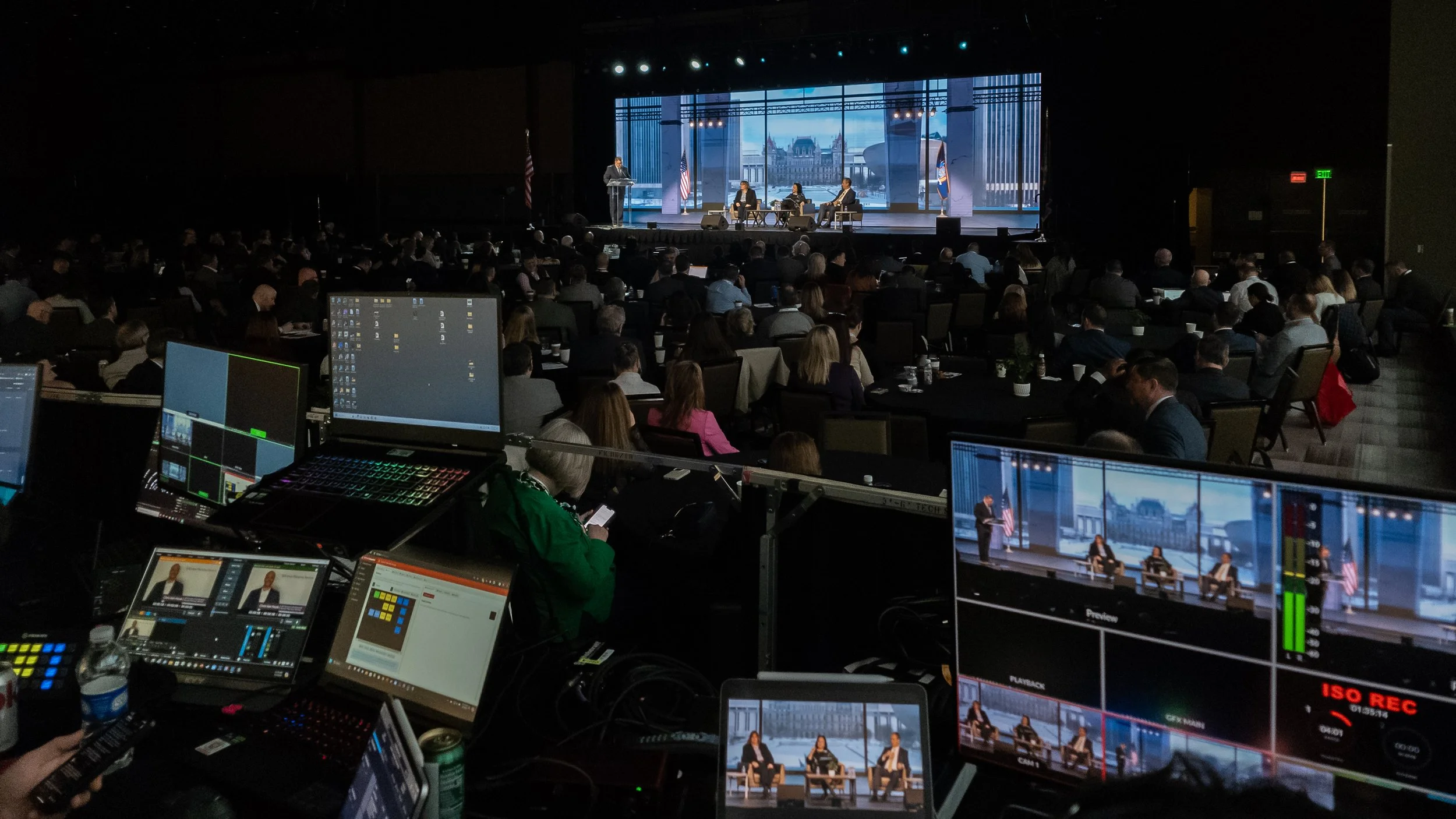 Audience watching a panel discussion or conference on stage with three seated speakers, with control monitors and production equipment in the foreground, and large screens displaying the event behind the stage.