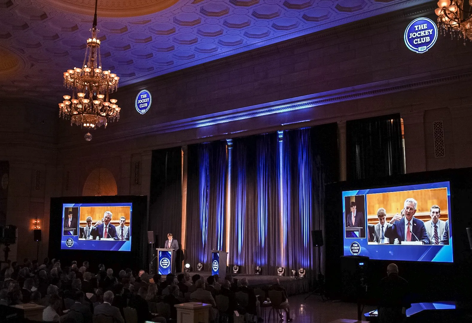 A speaker at a podium giving a speech during a conference or event at The Jockey Club, with two large screens displaying the speaker, in a large hall with chandeliers and audience seated facing the stage.