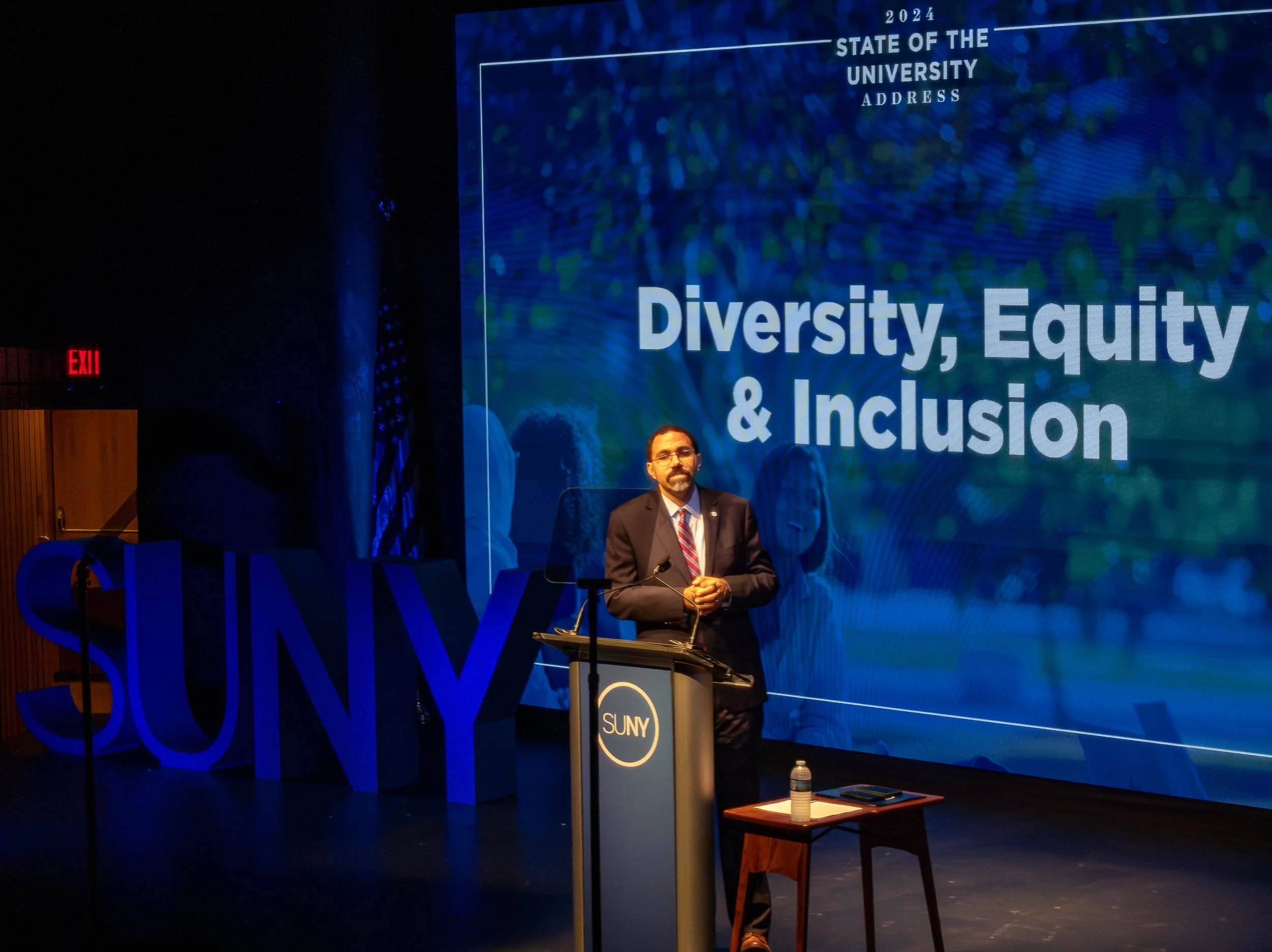 A man in a suit is standing at a podium on a stage, giving a presentation. Behind him is a large screen displaying the title 'Diversity, Equity & Inclusion' and the words 'State of the University Address' and '2024.' To his left, large blue letters s