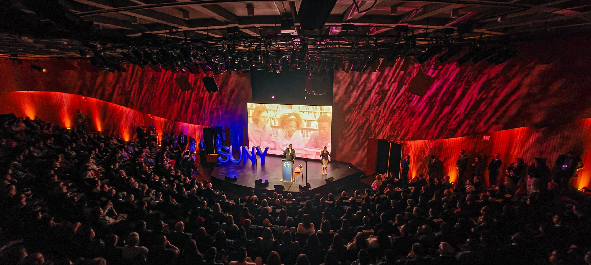 A large conference or presentation hall filled with an audience. On stage, a speaker stands at a podium, with a large screen behind displaying a video or presentation. The stage features the letters 'SUNY' in blue. The room is dimly lit with red and 