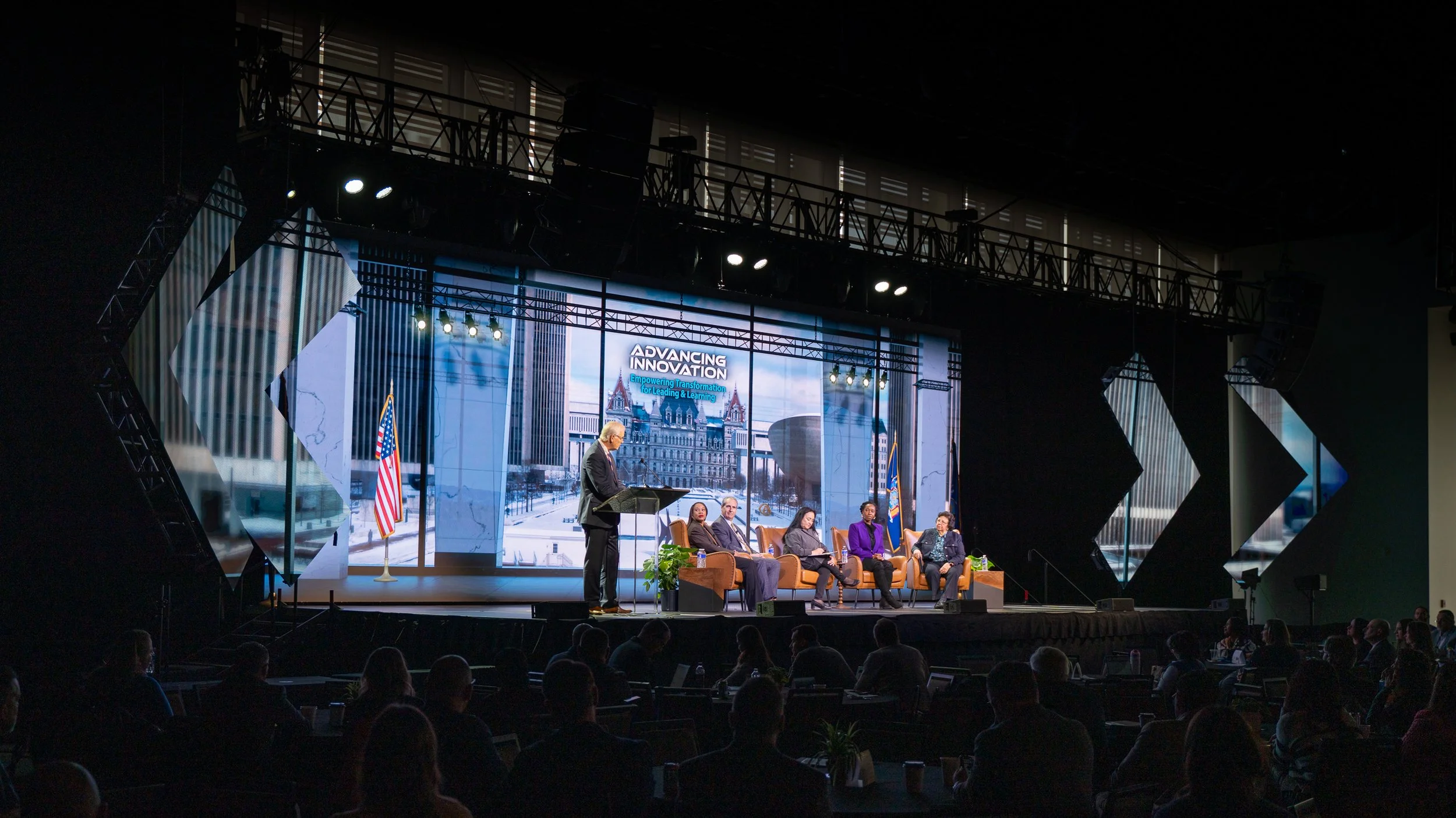 A conference or panel event on a stage with six people seated in chairs and one person standing at a podium, with a backdrop displaying 'Advancing Innovation' and an urban cityscape image, flags, and audience members watching.