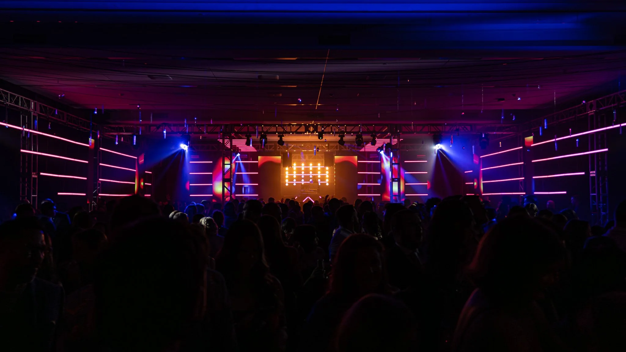 Darkened concert venue with a stage illuminated by vibrant pink, purple, and yellow lights, and a large crowd of silhouettes watching a DJ or performer.