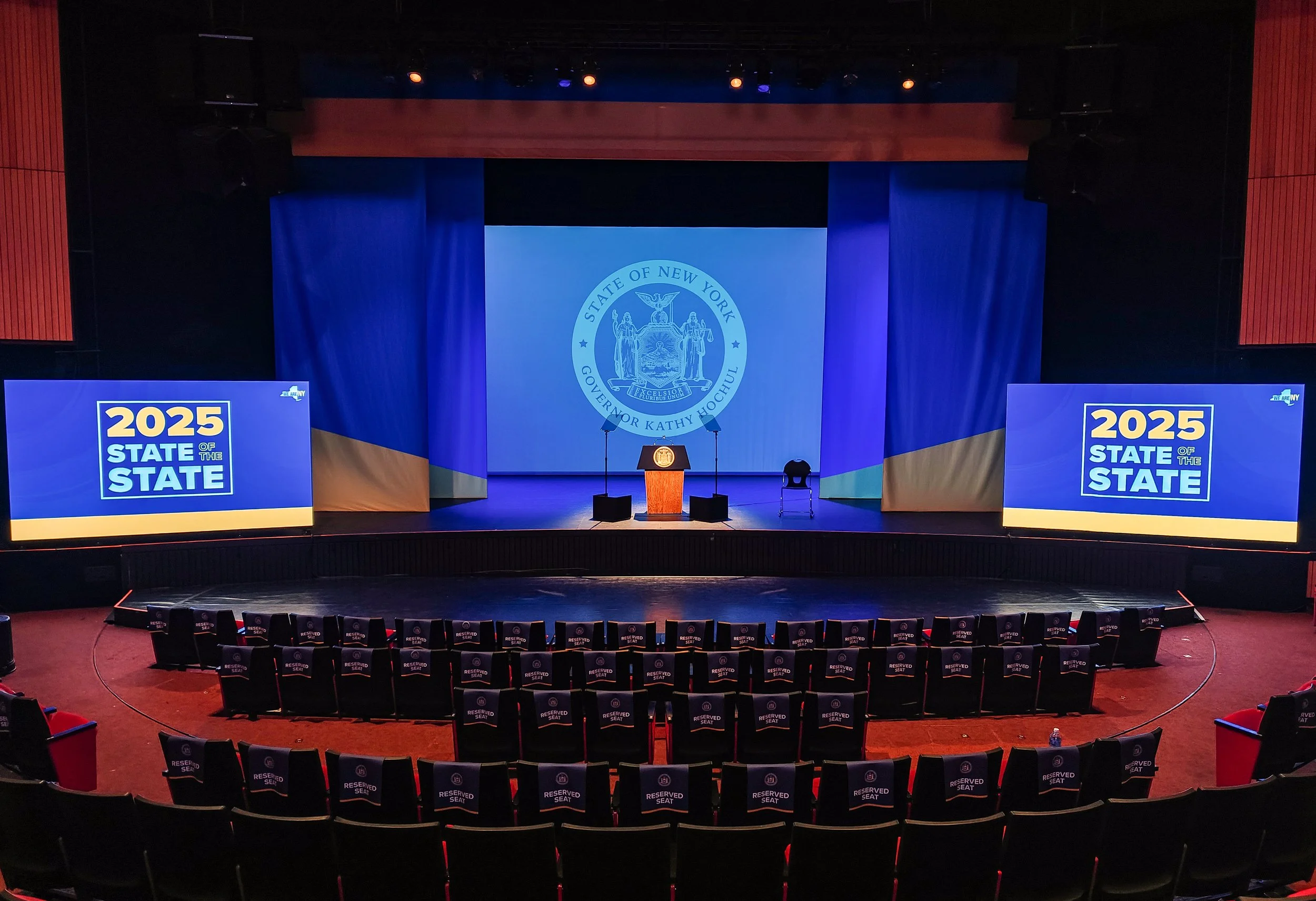 Empty stage with a podium in the center, flanked by two large screens displaying the 2025 State of the State website, and featuring the seal of the State of New York projected on the backdrop, with reserved seats in the foreground.