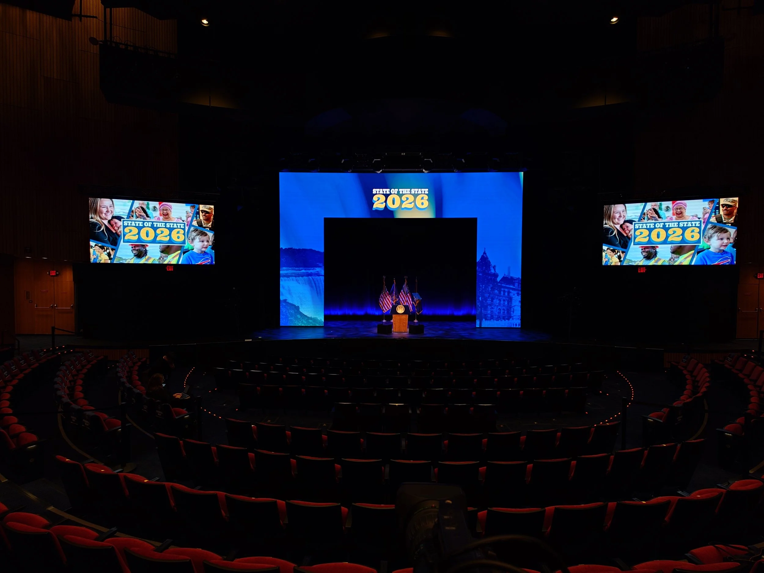 Empty auditorium with a stage at center featuring a podium and American flags, large screens on both sides display a collage of diverse people and the text "State of the State 2026", and a blue backdrop with a waterfall image.