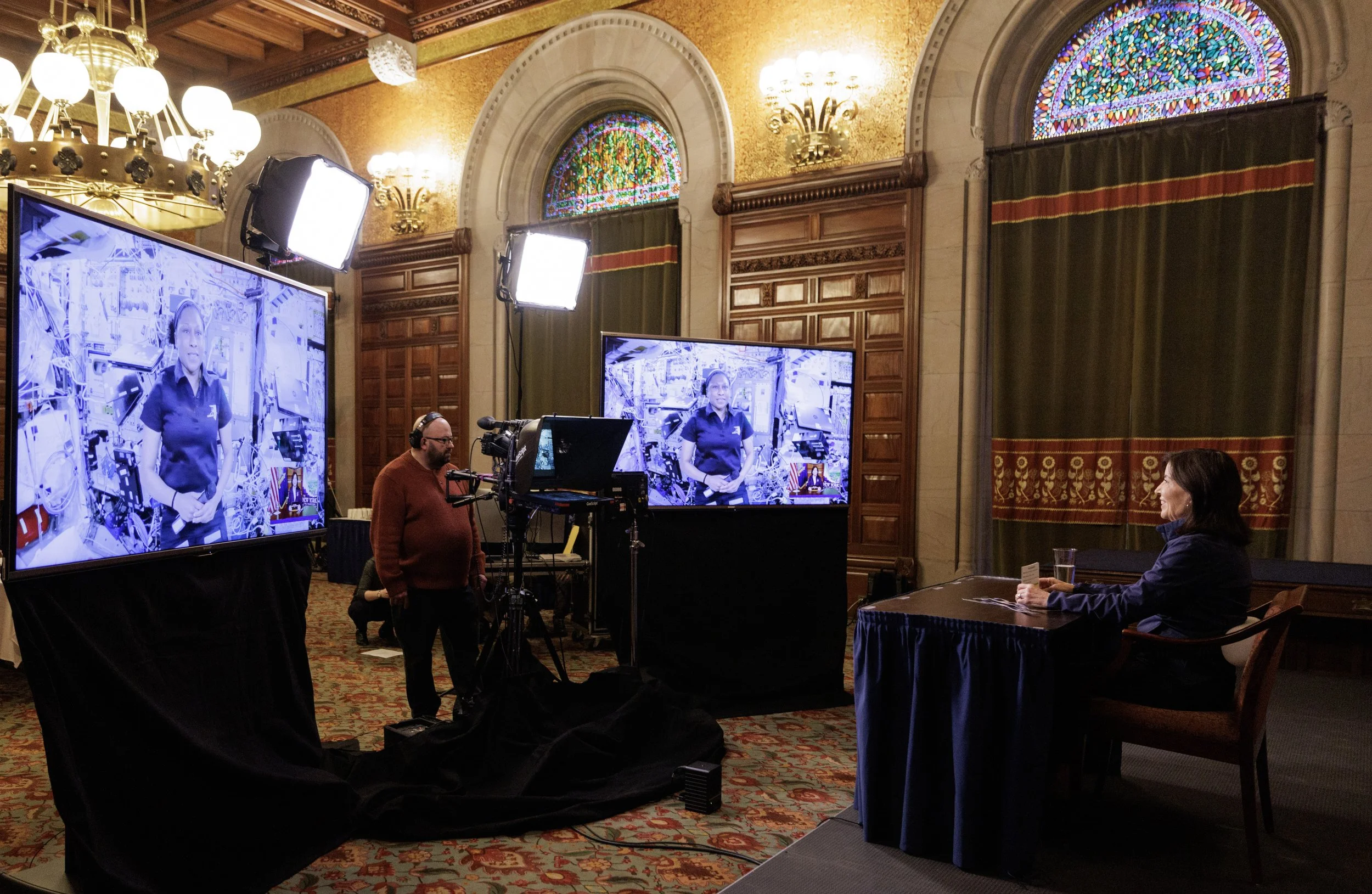 A woman in a dark jacket sitting at a table in a grand, ornate room with stained glass windows, participating in a virtual meeting or interview. There are two large screens showing her image, surrounded by professional lighting and camera equipment.