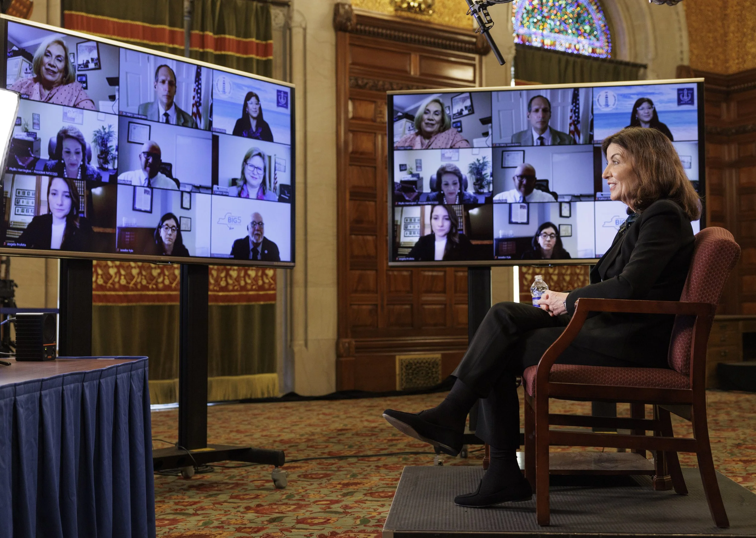 A woman sitting on a chair holding a water bottle during a virtual conference with multiple screens showing participants in a formal setting with wood paneling, chandeliers, and a stained glass window.