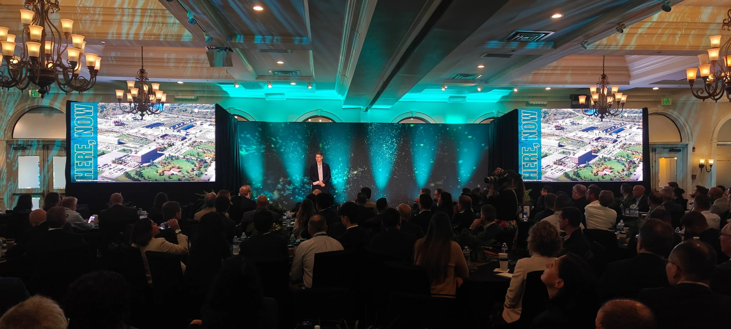 A man is speaking on stage at a conference, with a large screen displaying an aerial view of a business complex behind him. The room is filled with seated attendees. The stage has a backdrop with teal lighting and decorative sparks.