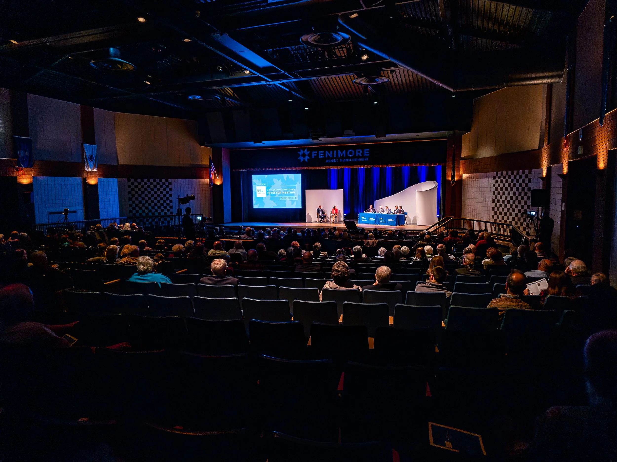 An audience attending a conference in a large auditorium with a stage, a presentation screen, and panelists seated at a table.