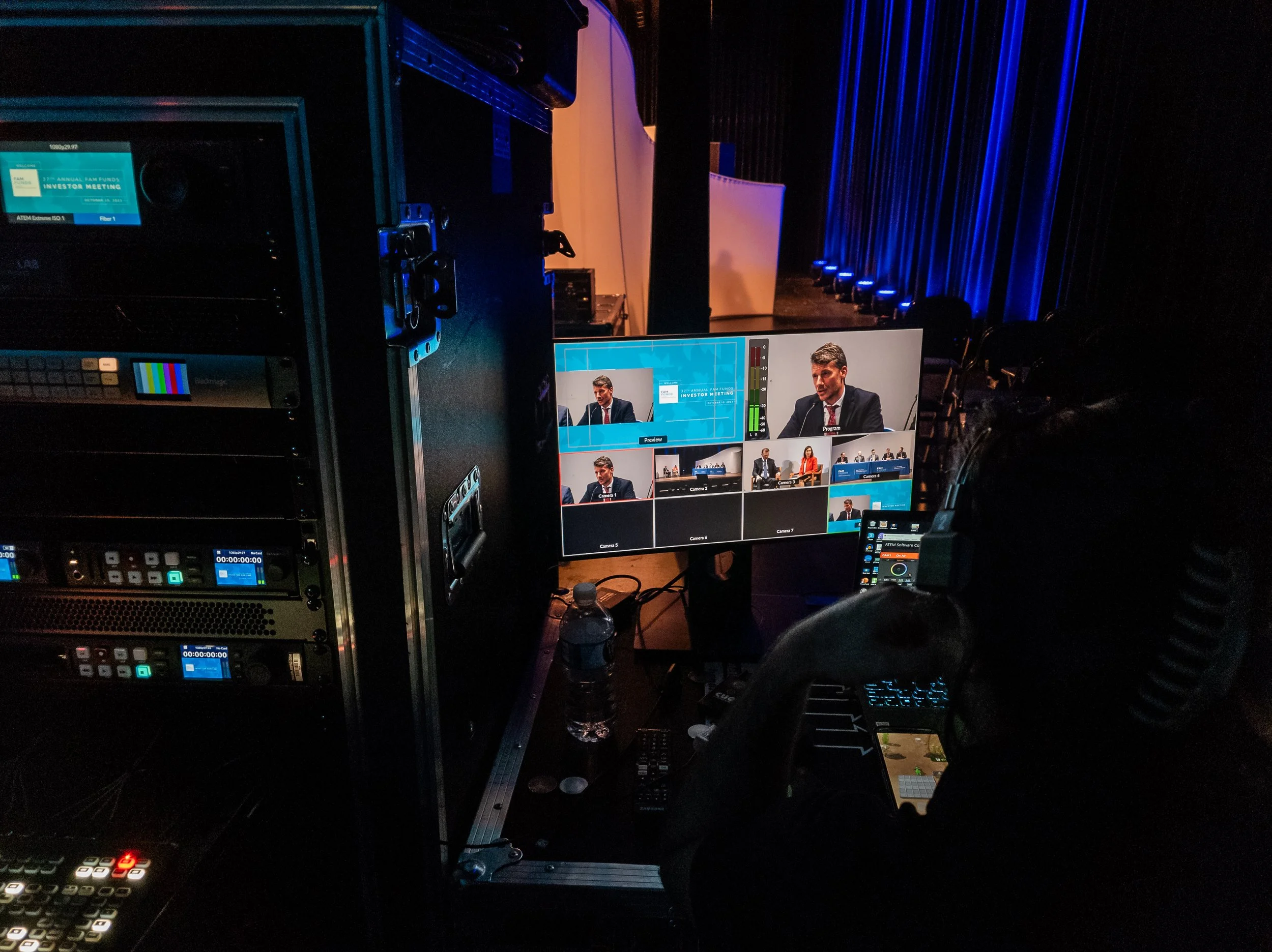 A person wearing headphones operating a video switcher and a computer with multiple monitors showing a live video feed of a panel discussion on stage with several panelists, some seated at a table, in a dimly lit auditorium with blue stage curtains.