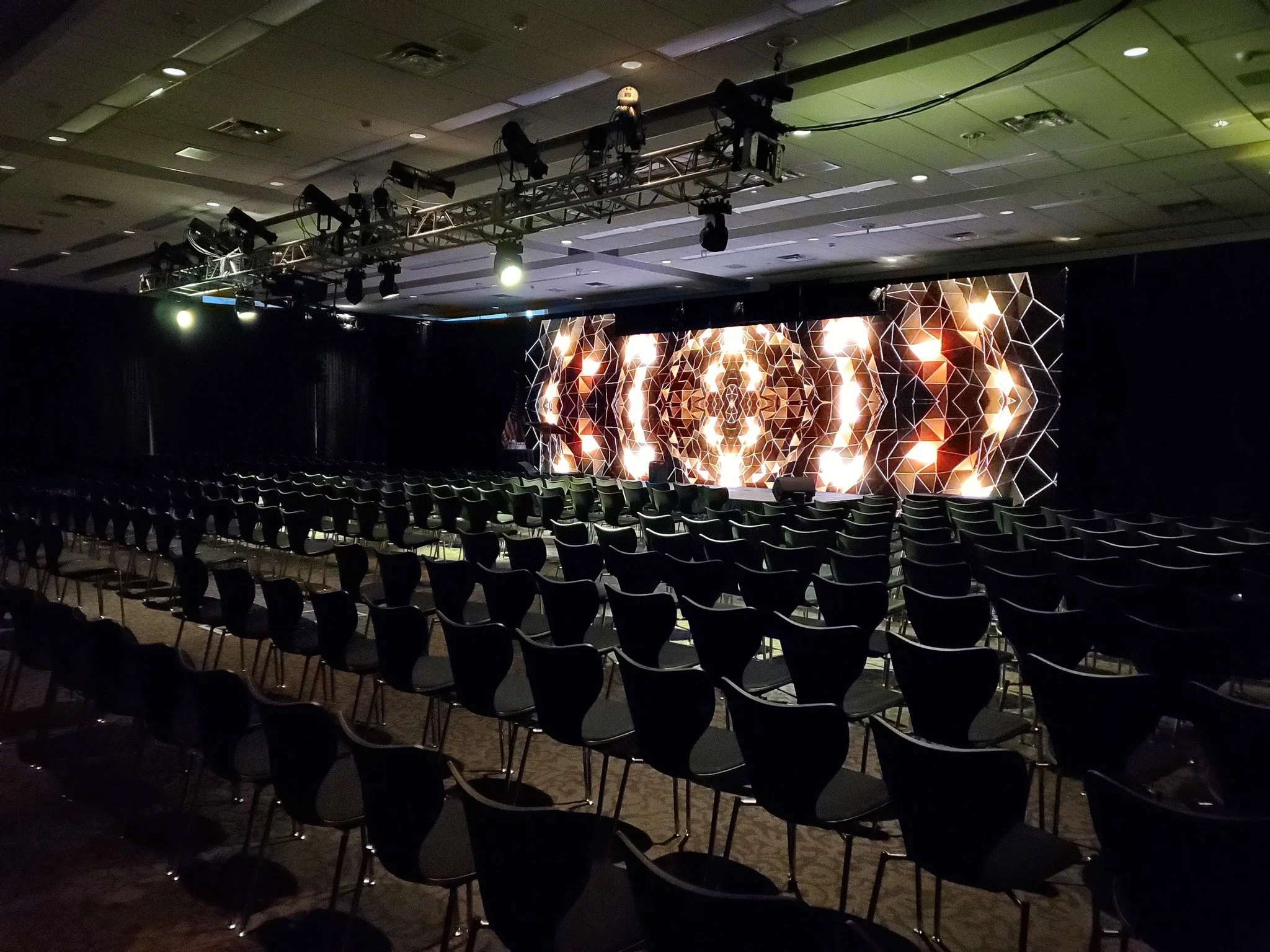 Empty conference room with rows of black chairs facing a stage with a large digital display showing abstract geometric patterns and lighting effects.