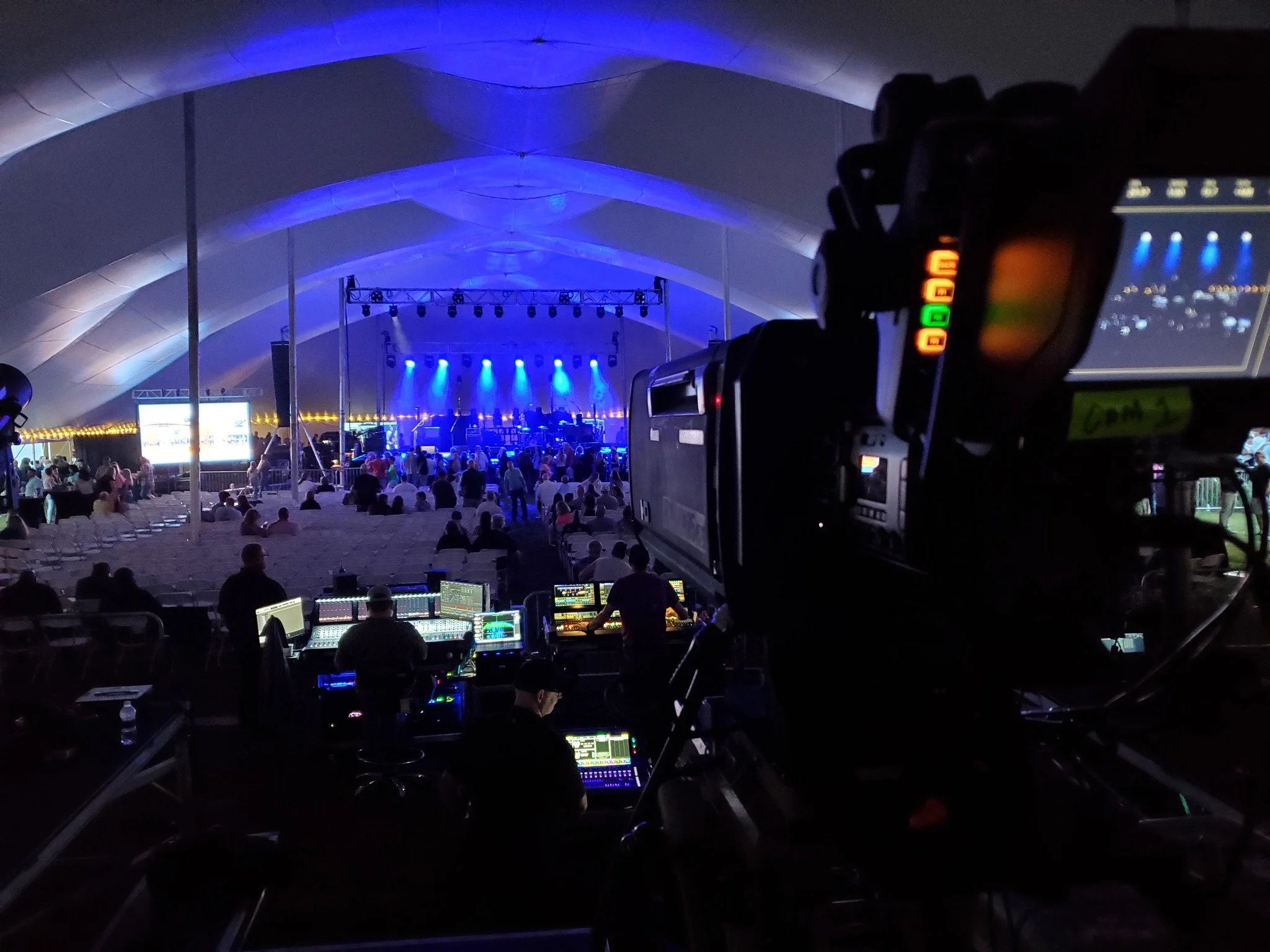 View of a stage with blue lighting at a concert or event, seen from behind a camera and mixing console, with an audience seated under a white tent canopy.