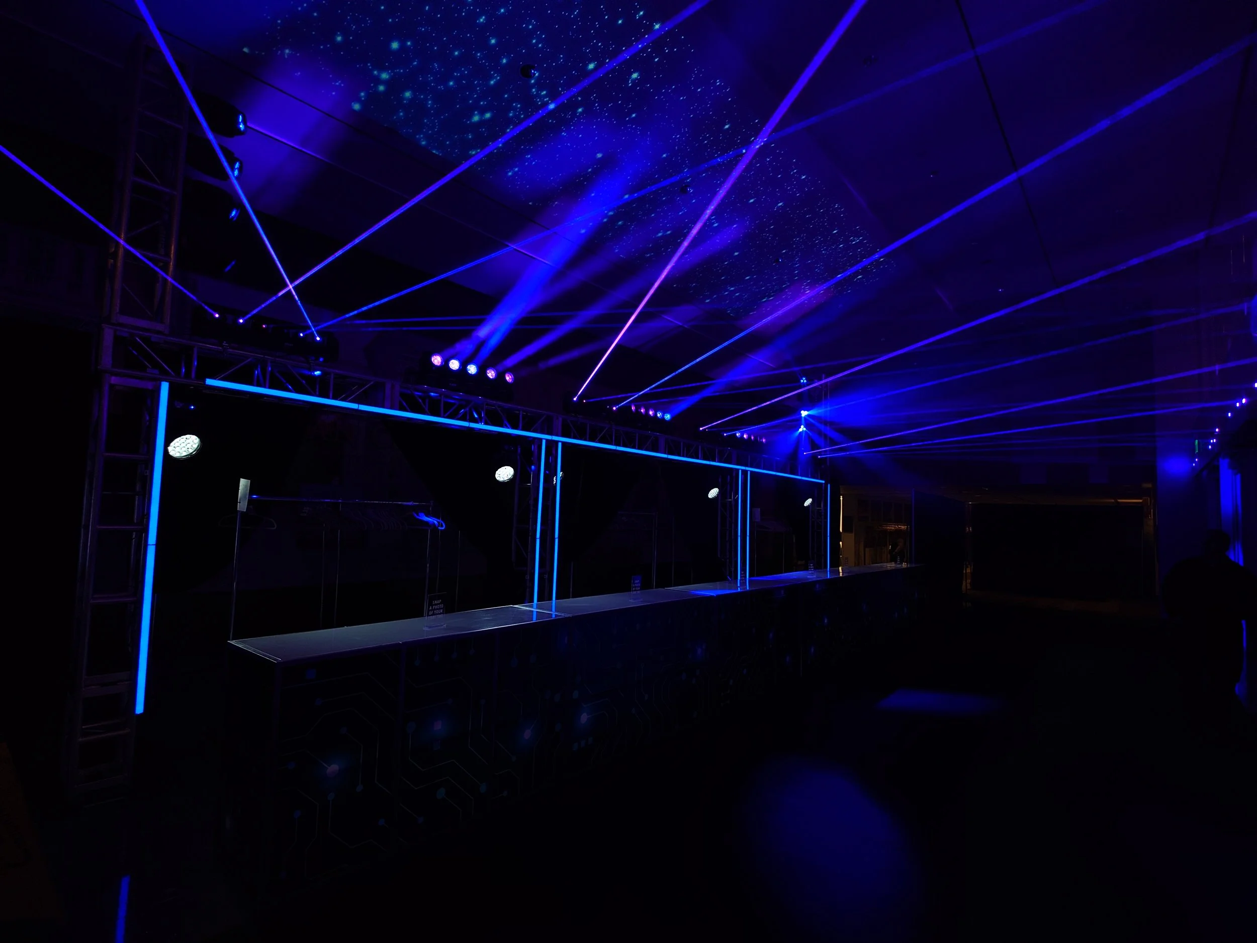 Dark room with blue and purple laser lights creating a starry effect on the ceiling and illuminating a thin, reflective surface, possibly a bar or display counter, with a metal frame structure.