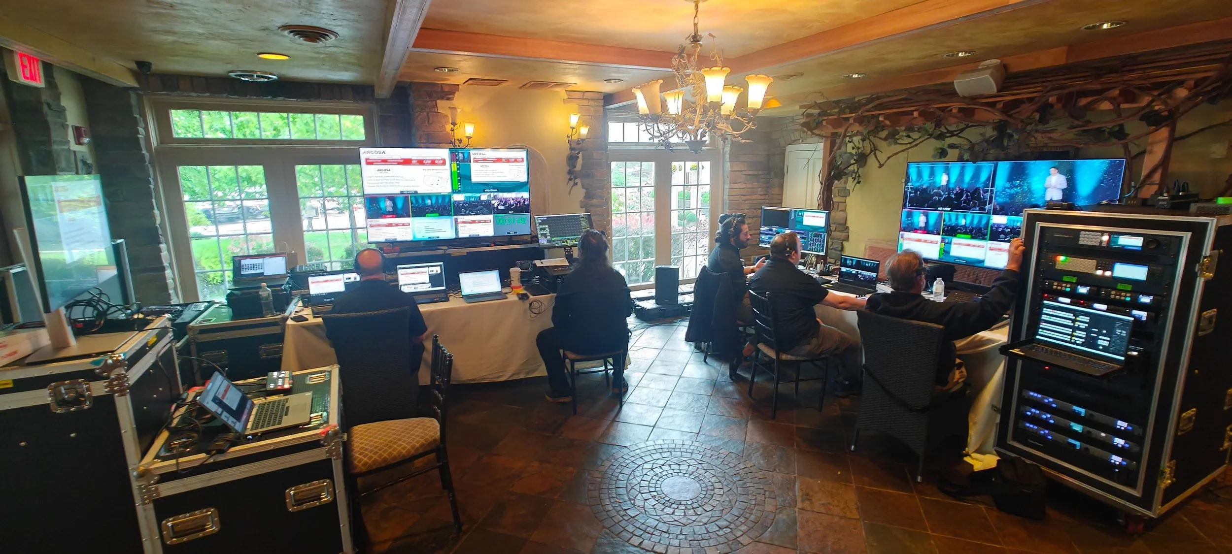 A behind-the-scenes view of a broadcast control room with multiple operators working at their stations with large monitors, audio equipment, and a chandelier hanging from a rustic ceiling, in a warmly lit room with windows and visible outdoor greener