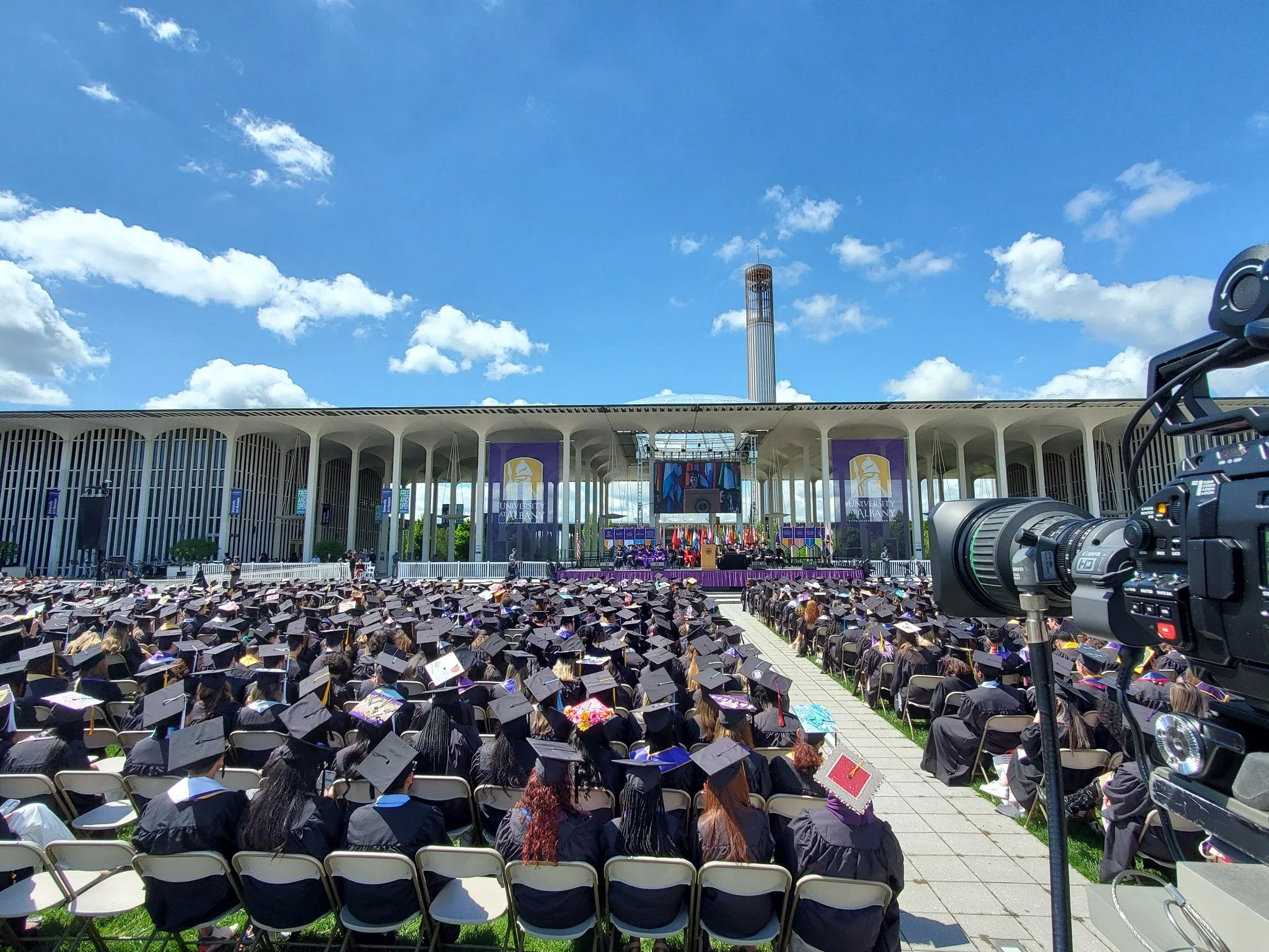 A large outdoor graduation ceremony with students in caps and gowns seated in rows, facing a stage with a large screen and banners at a modern architectural building under a blue sky with clouds.