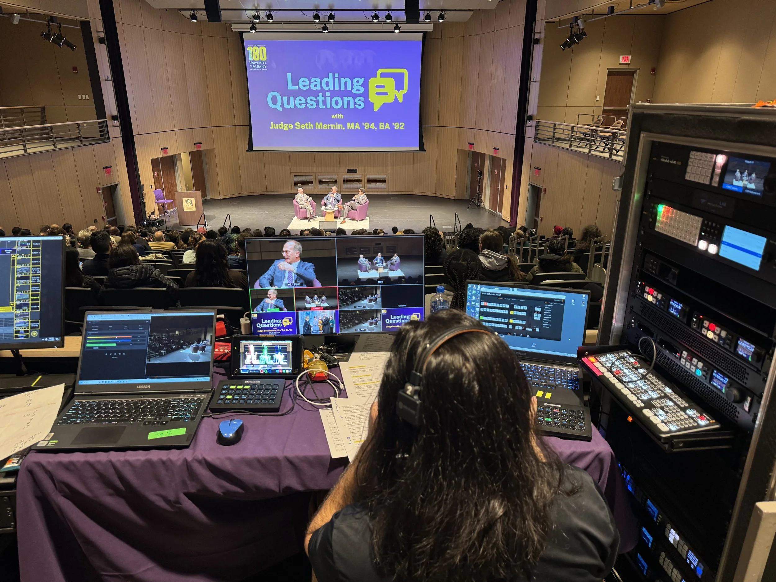Stage with three people in conversation and a large screen behind them displaying text about a discussion with Judge Seth Marnin at an auditorium. Audience seated facing the stage. Technical equipment and operator in foreground setup for live streami