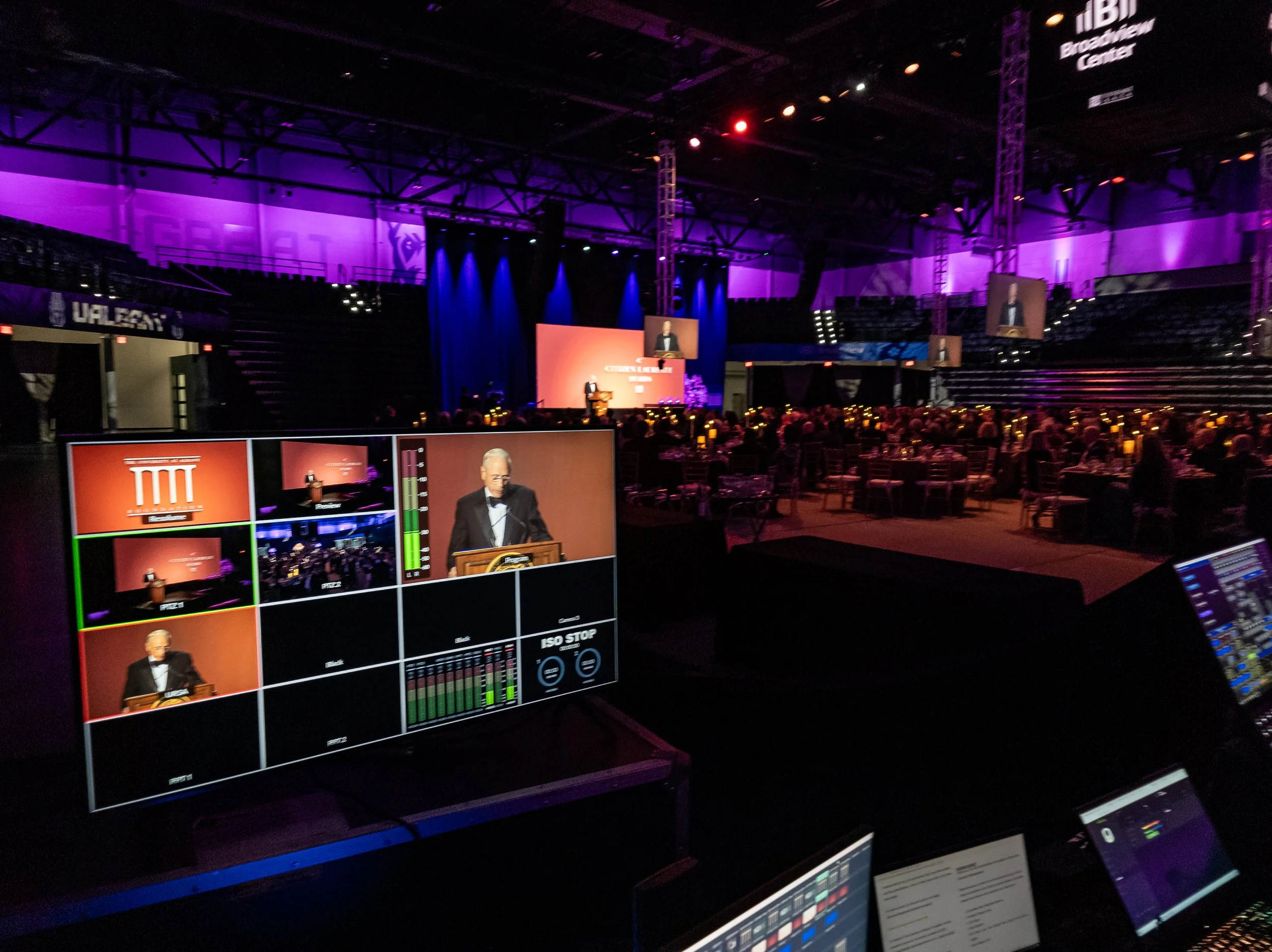 An event stage with a speaker at a podium is visible on a large screen, with purple lighting and seating for an audience in an indoor arena.