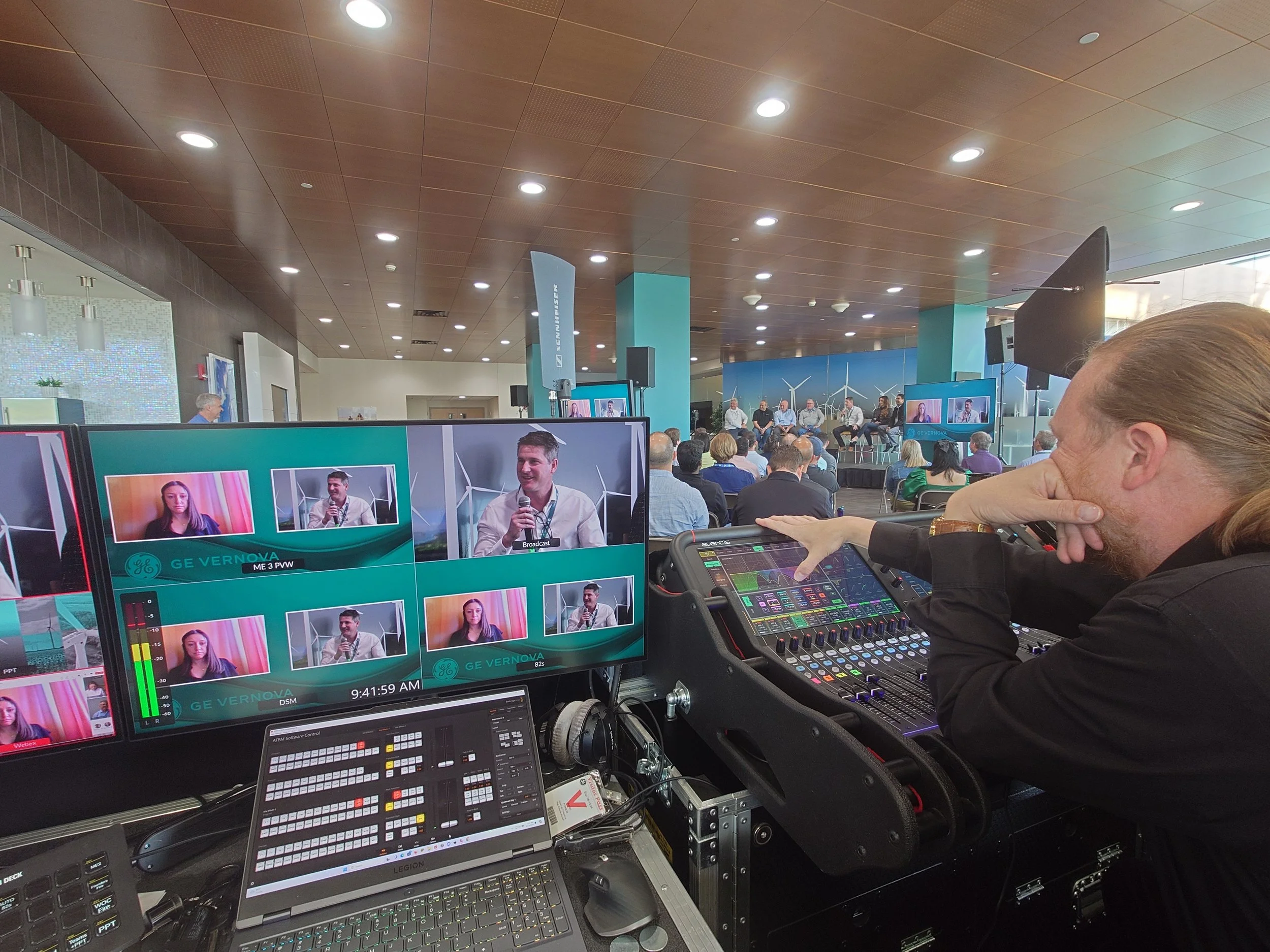 A man operating a video control panel during a conference with a panel discussion on stage in the background.
