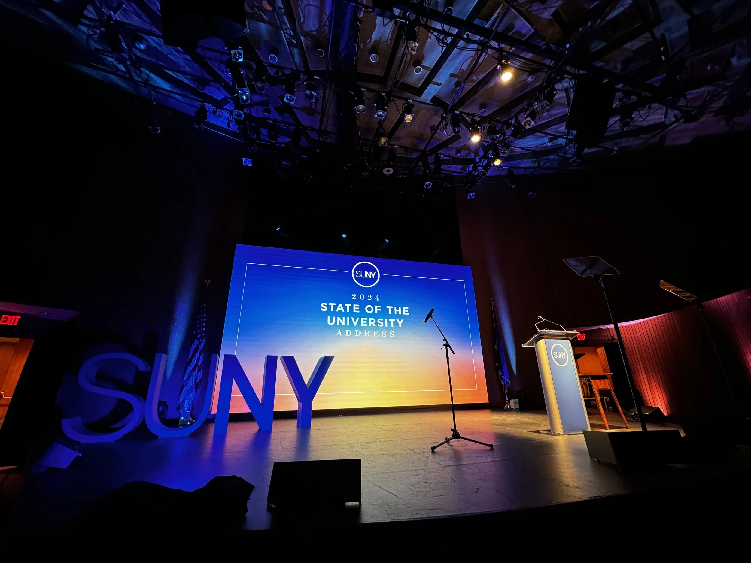 Stage set for SUNY 2024 State of the University address, with large SUNY letters on the left and a podium on the right, in front of a large digital screen displaying the event details and a sunset background.
