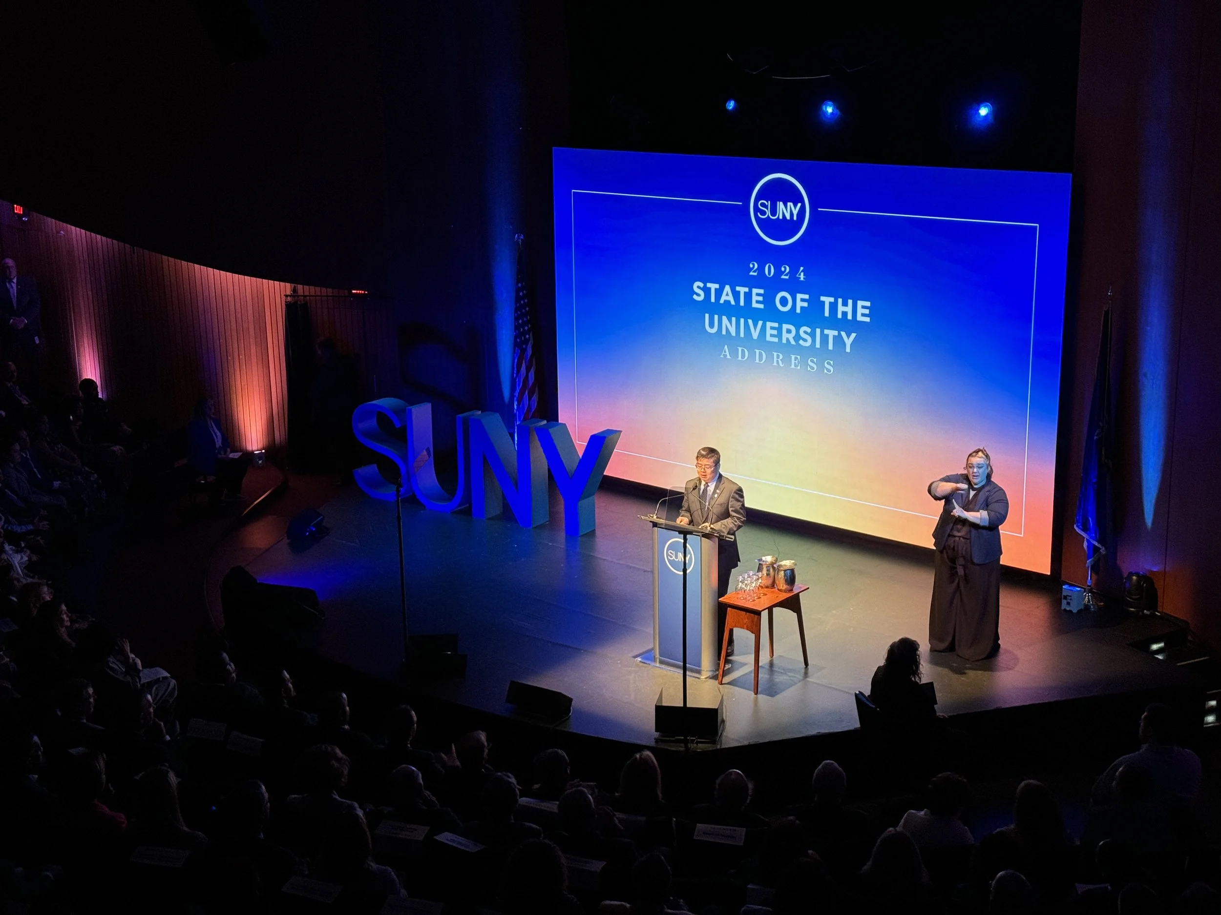A speaker on stage at a SUNY event titled '2024 State of the University Address.' A large screen behind shows the event details, with an audience seated in front.