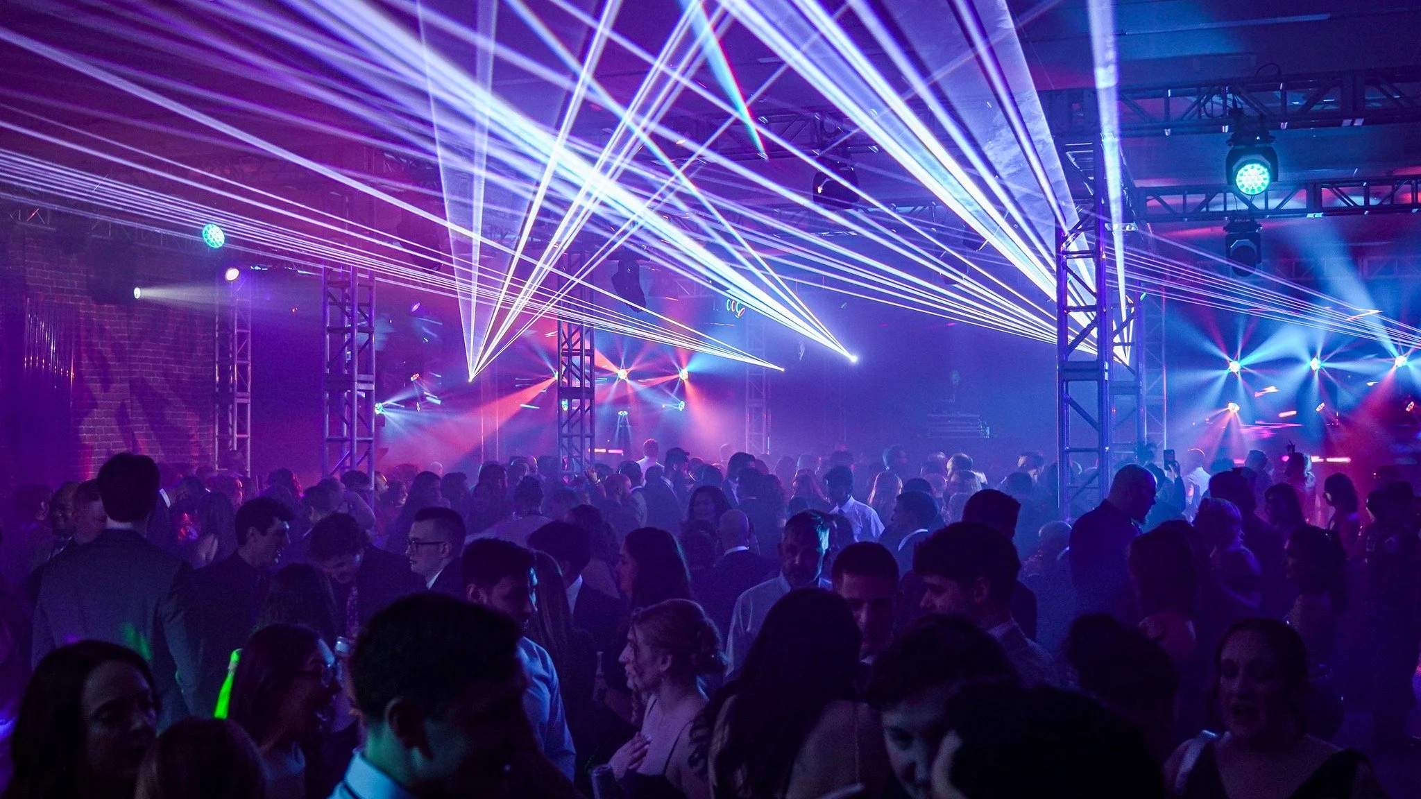 A crowded dance party with purple and blue laser lights, people dancing and socializing, in a nightclub or event space.